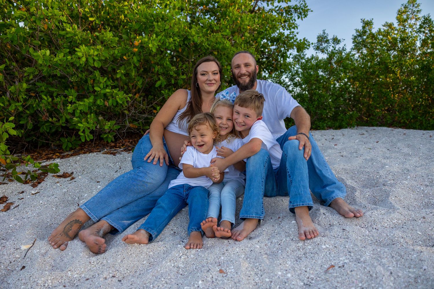 Happy family of five posing together on a sandy beach with lush green trees in the background.