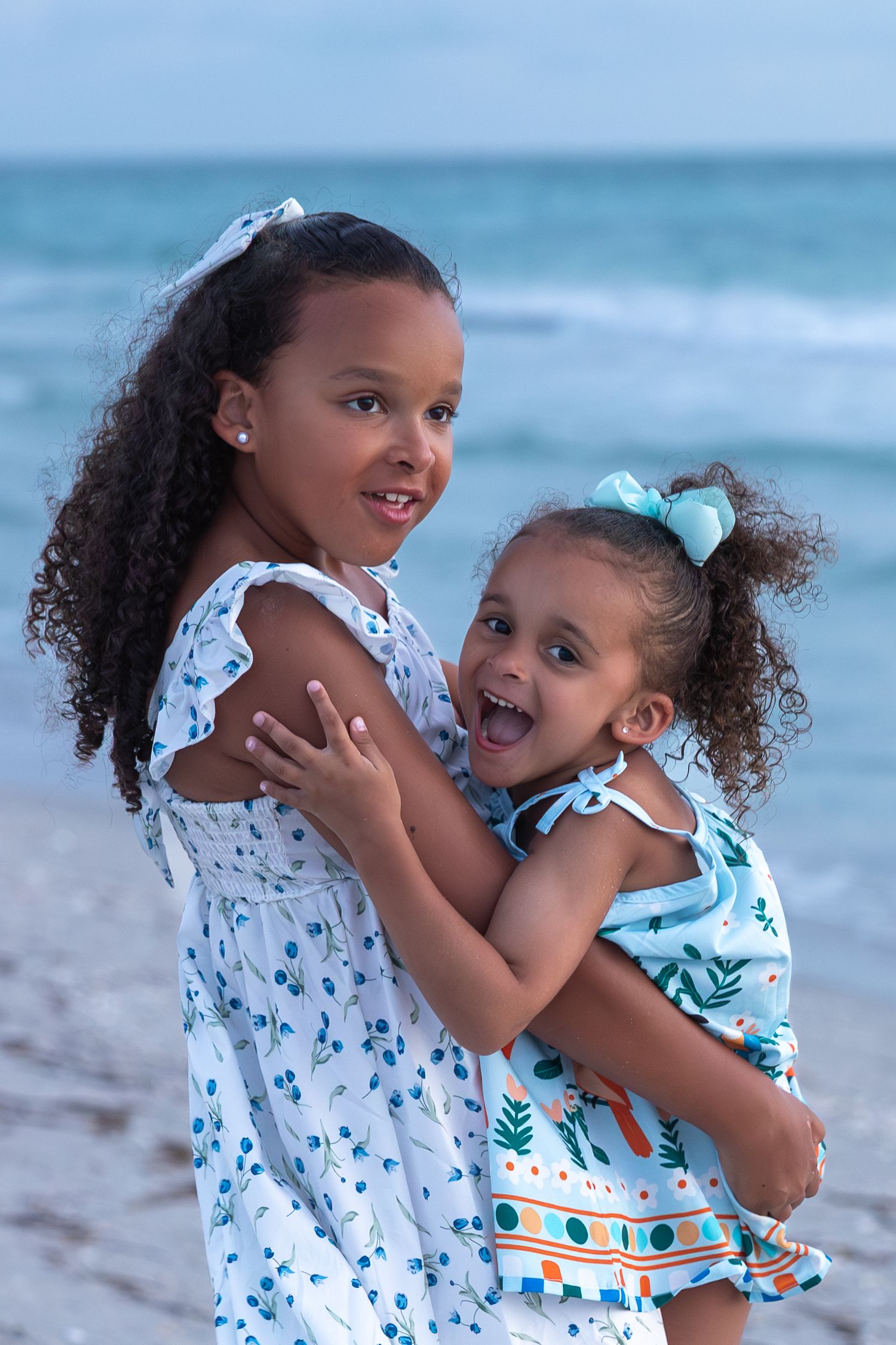 Two young sisters in matching floral dresses hugging and smiling on a beach with ocean waves in the background.