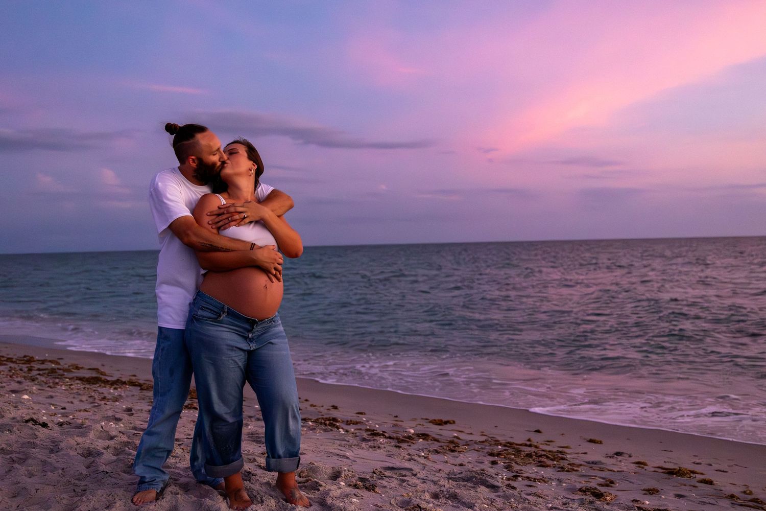 Pregnant couple embracing on a beach at sunset with pink and purple sky over the ocean.