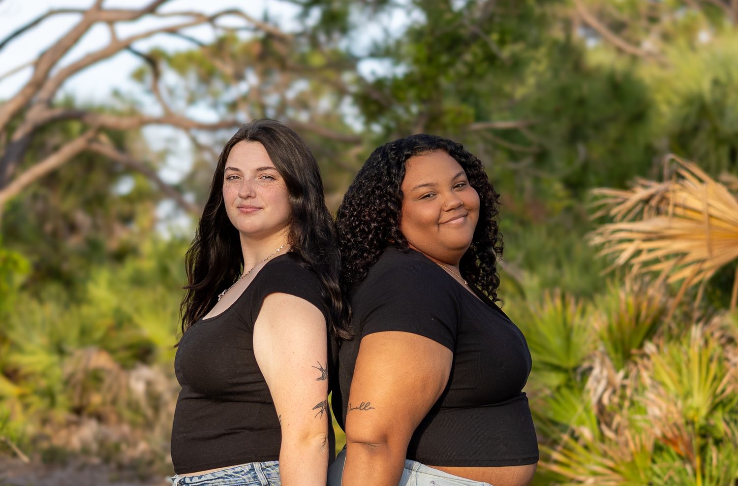 Two young women standing back to back smiling outdoors among lush green trees.