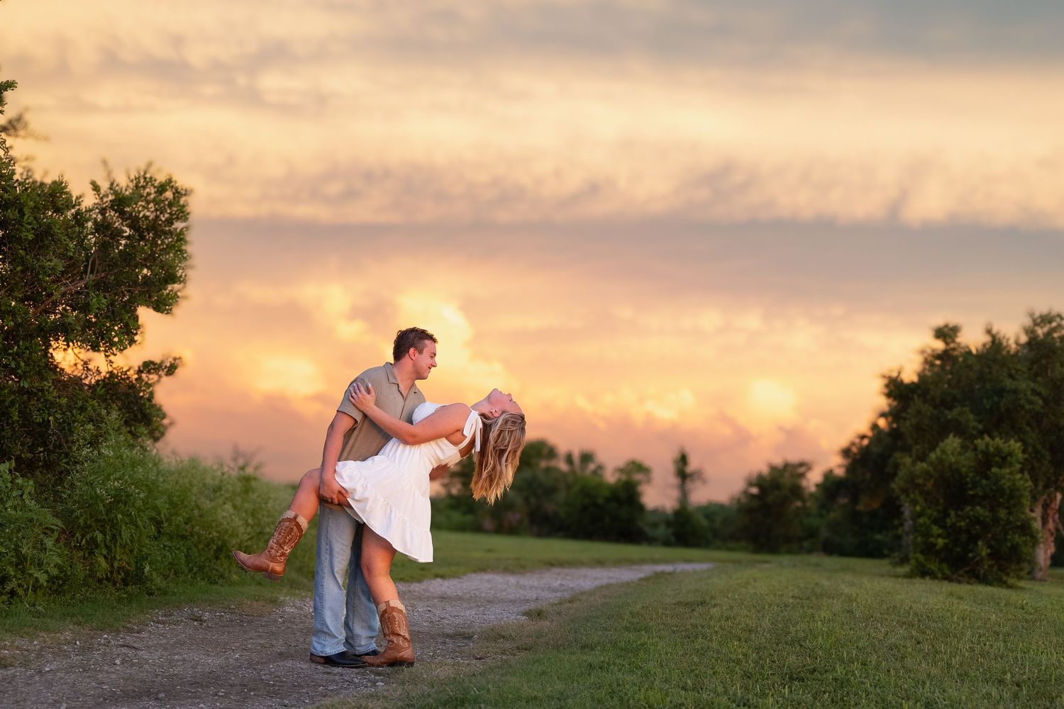 Couple sharing a romantic dip kiss on a country path at golden sunset, surrounded by lush green trees.