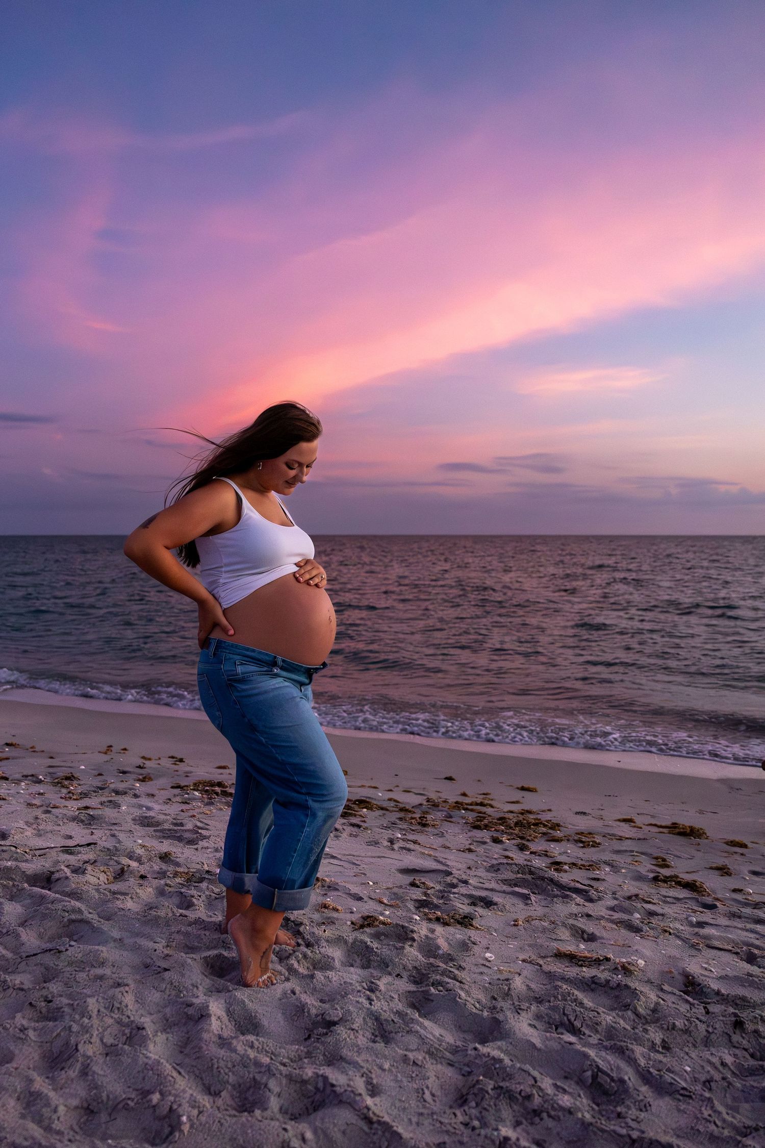 Pregnant woman in white top and jeans cradling her baby bump on a sandy beach at a vibrant pink and purple sunset.