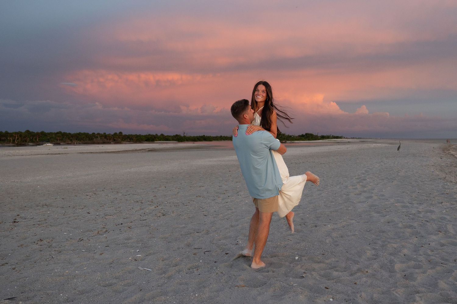 Couple embracing on sandy beach at sunset with vibrant pink and purple clouds in the background.