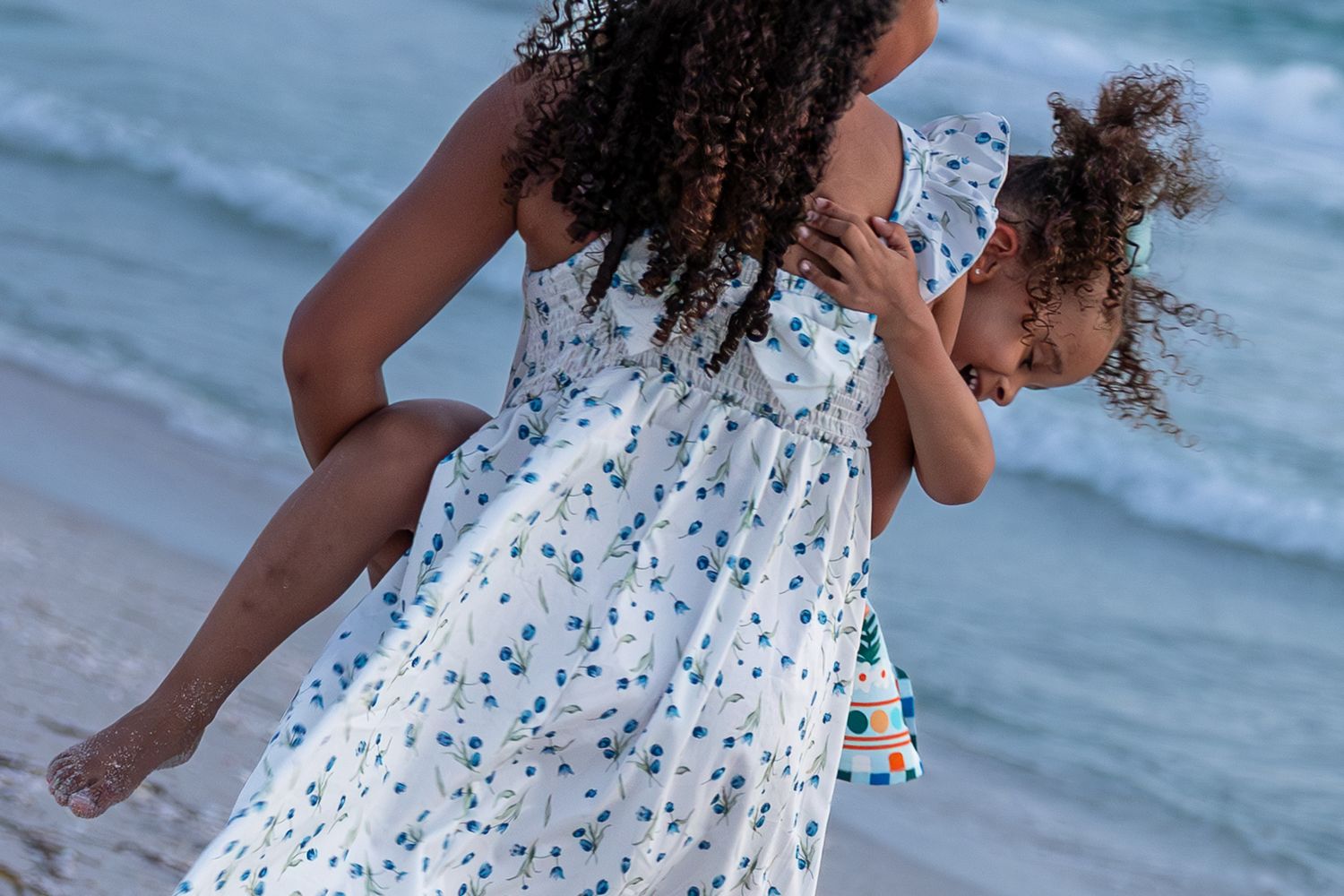 A mother in a floral dress carries her young daughter on a sunny beach near ocean waves.