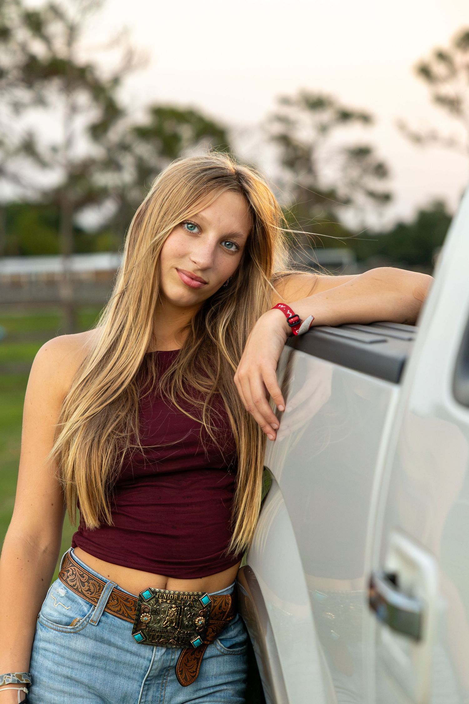 Young blonde woman in maroon crop top and jeans leaning against a white truck outdoors at sunset.