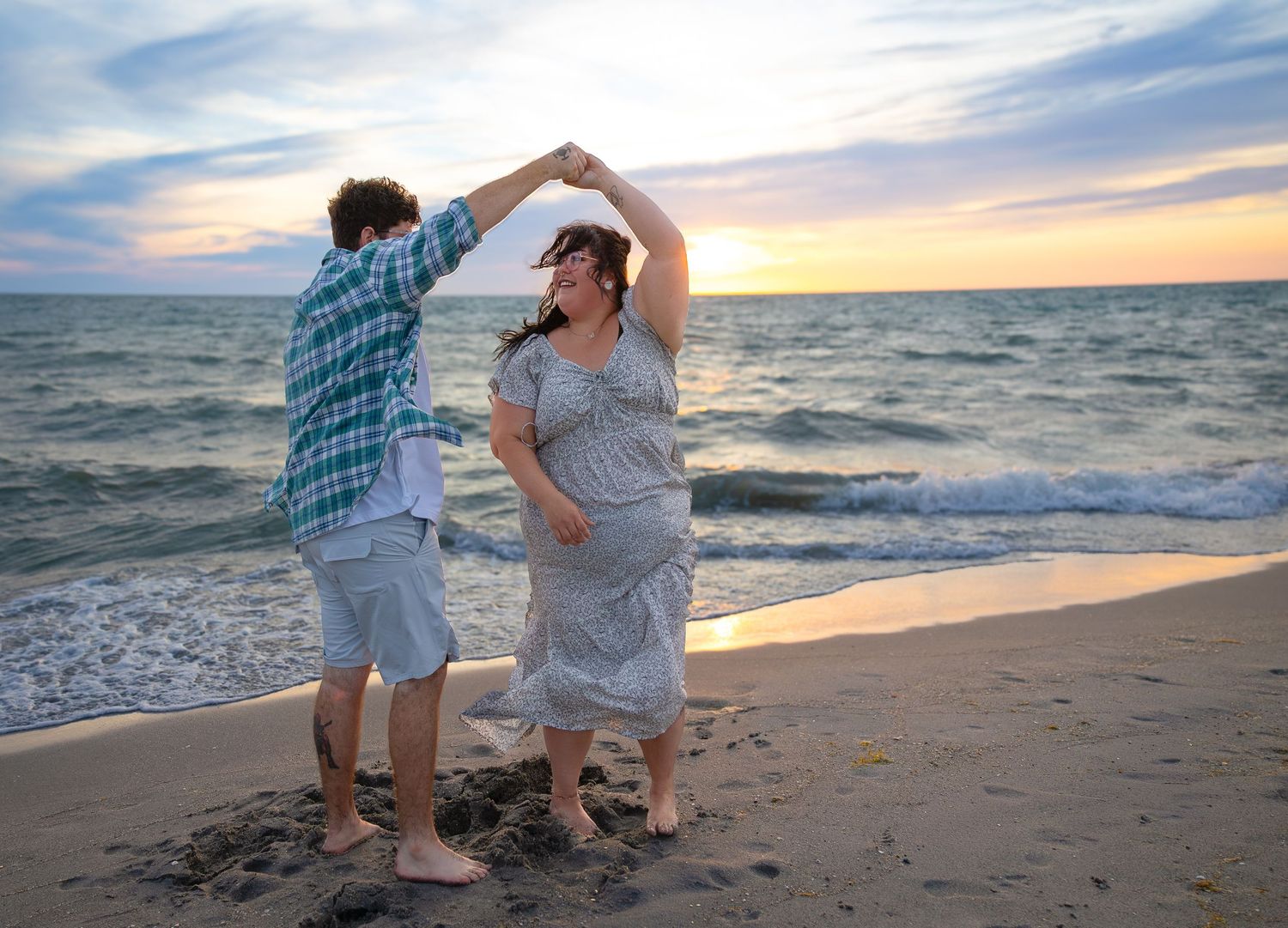 Couple dancing joyfully on a beach at sunset, woman in floral dress being twirled by partner.