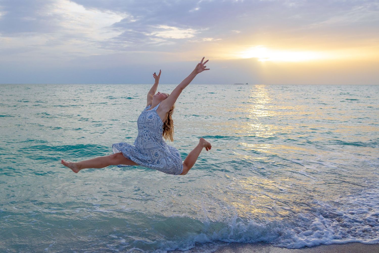 A joyful woman in a white dress leaps gracefully on a beach at sunset, arms raised against a colorful ocean sky.