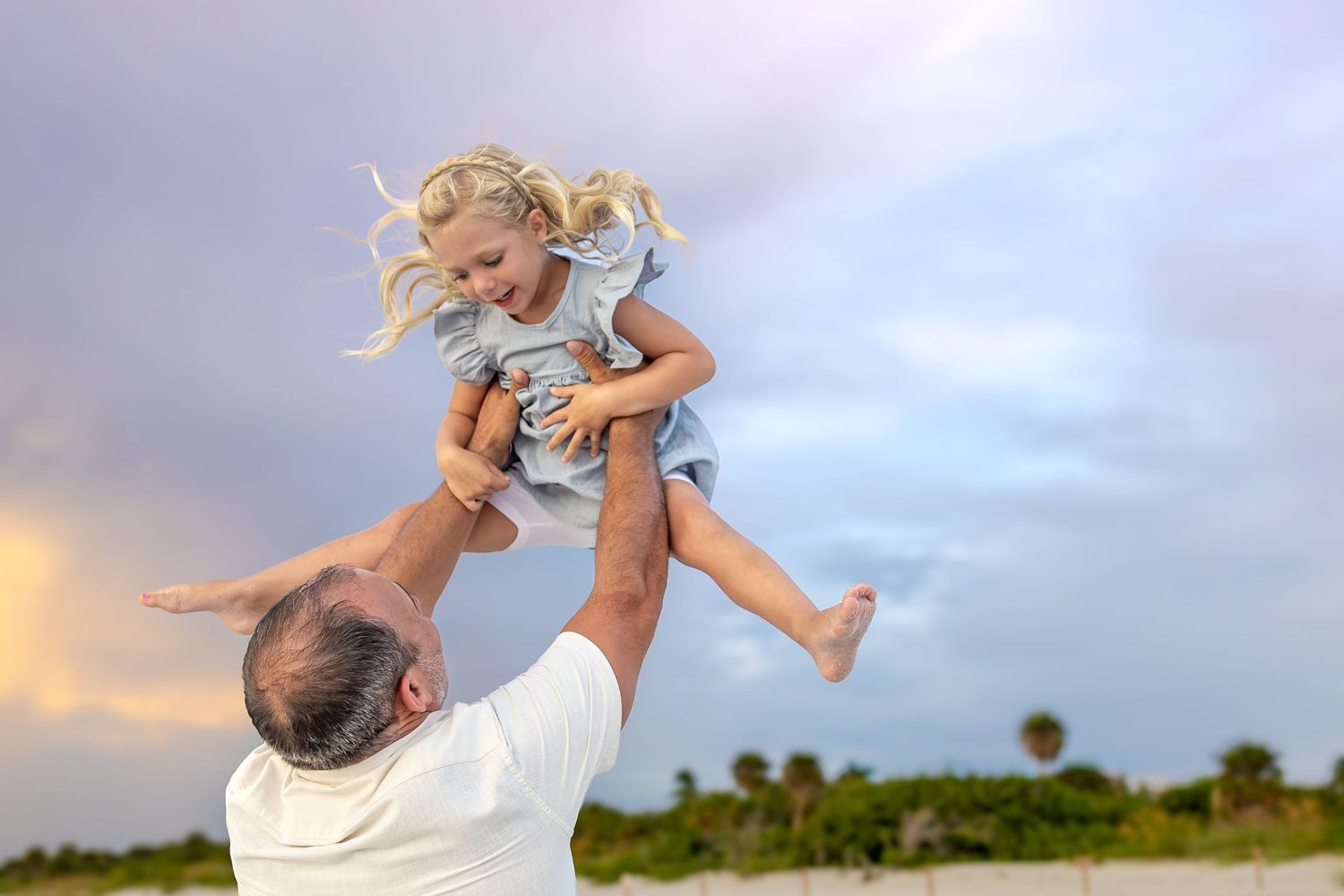 Father joyfully lifting smiling blonde girl in blue dress on beach at sunset with cloudy sky.