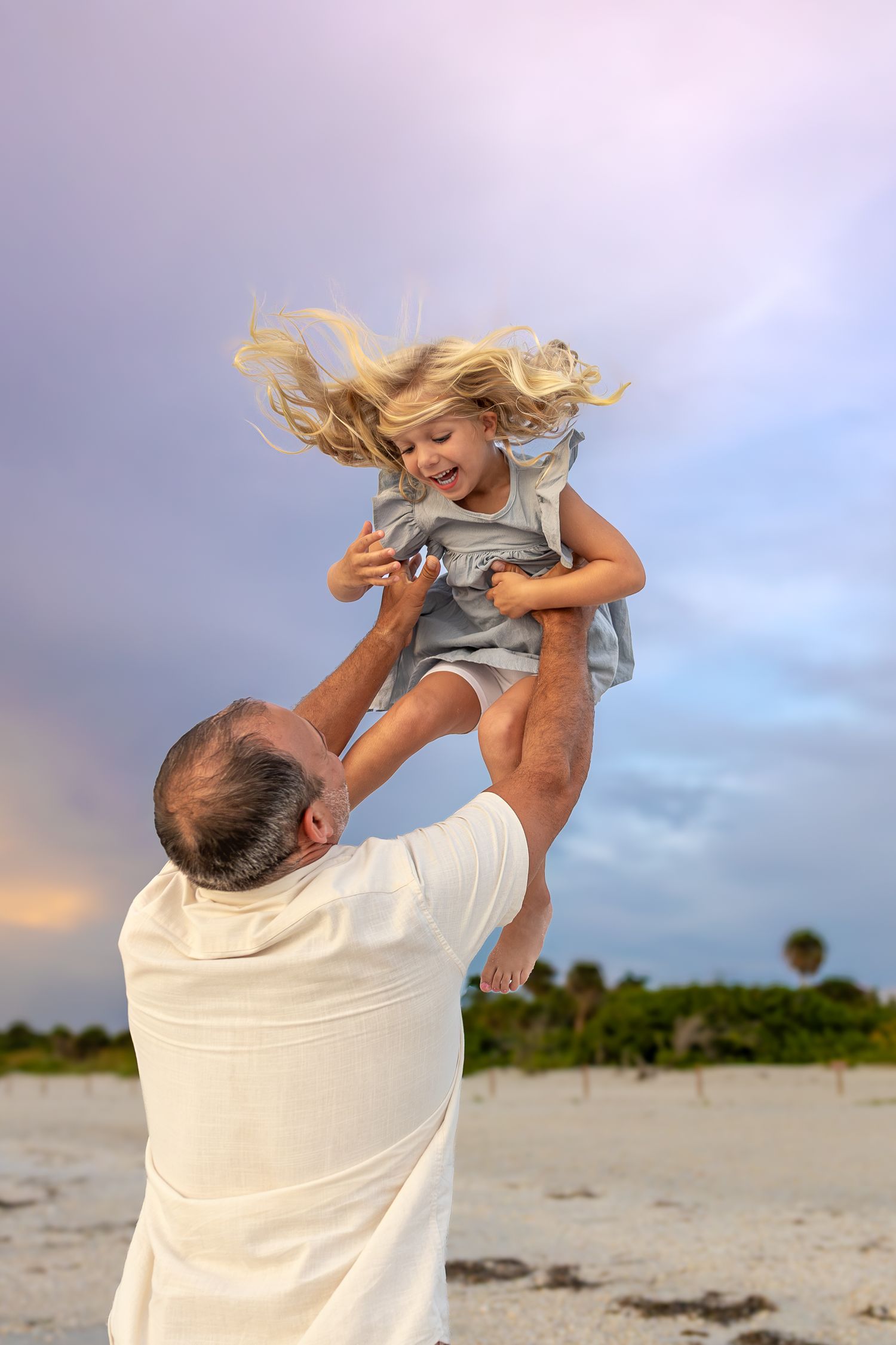 A joyful father lifting his laughing blonde daughter into the air on a sandy beach at sunset.