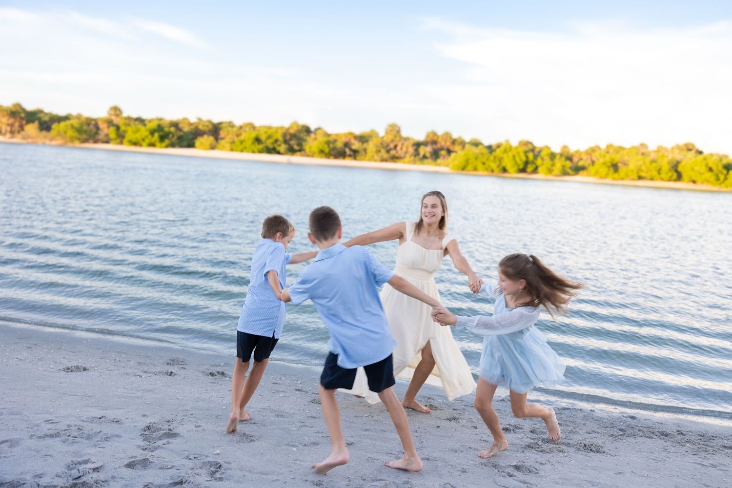 A joyful mother in a white dress plays with three children on a sunny beach near calm water and lush green trees.