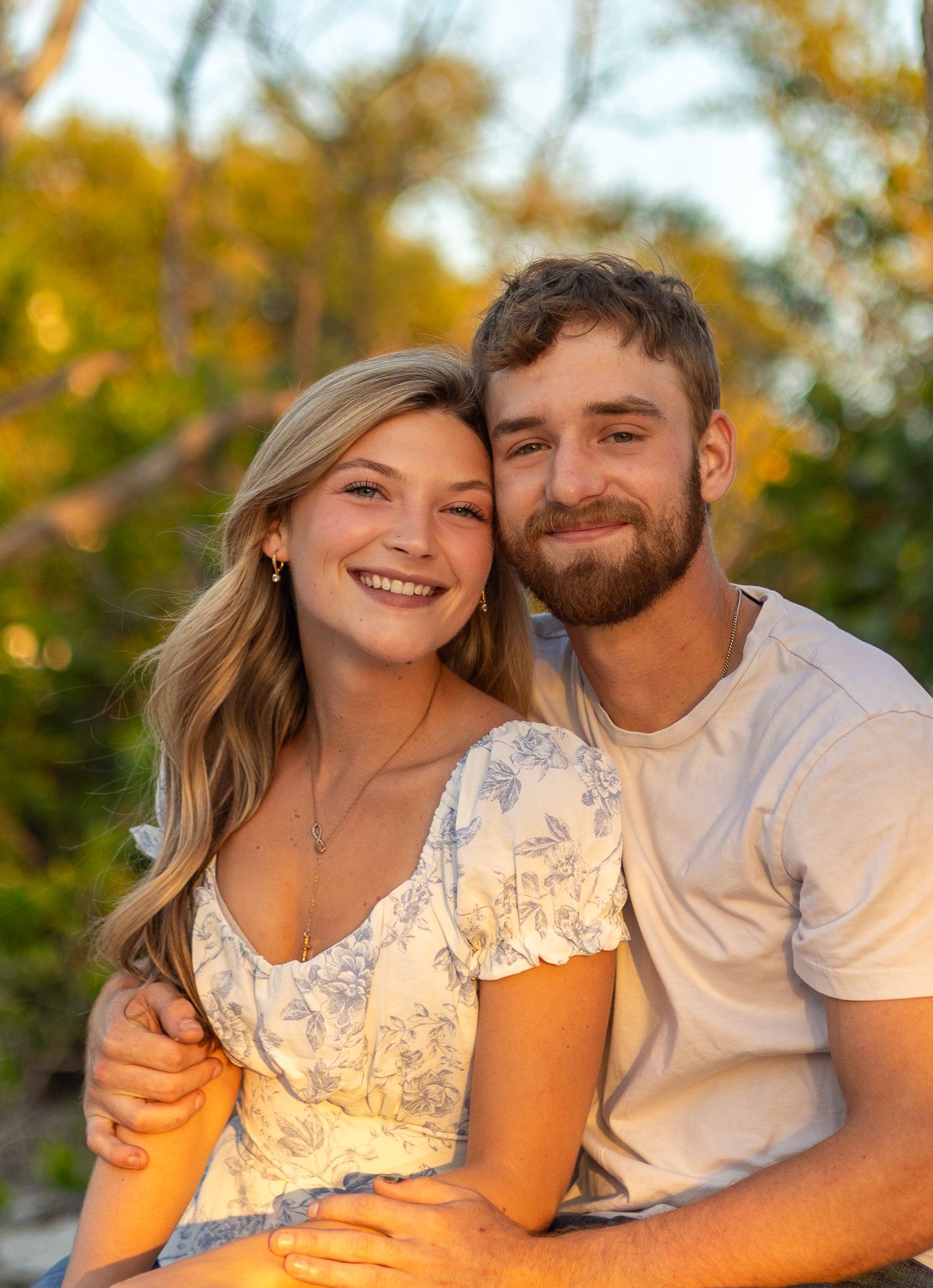 A joyful young couple poses together outdoors with colorful fall trees in the warm sunset background.
