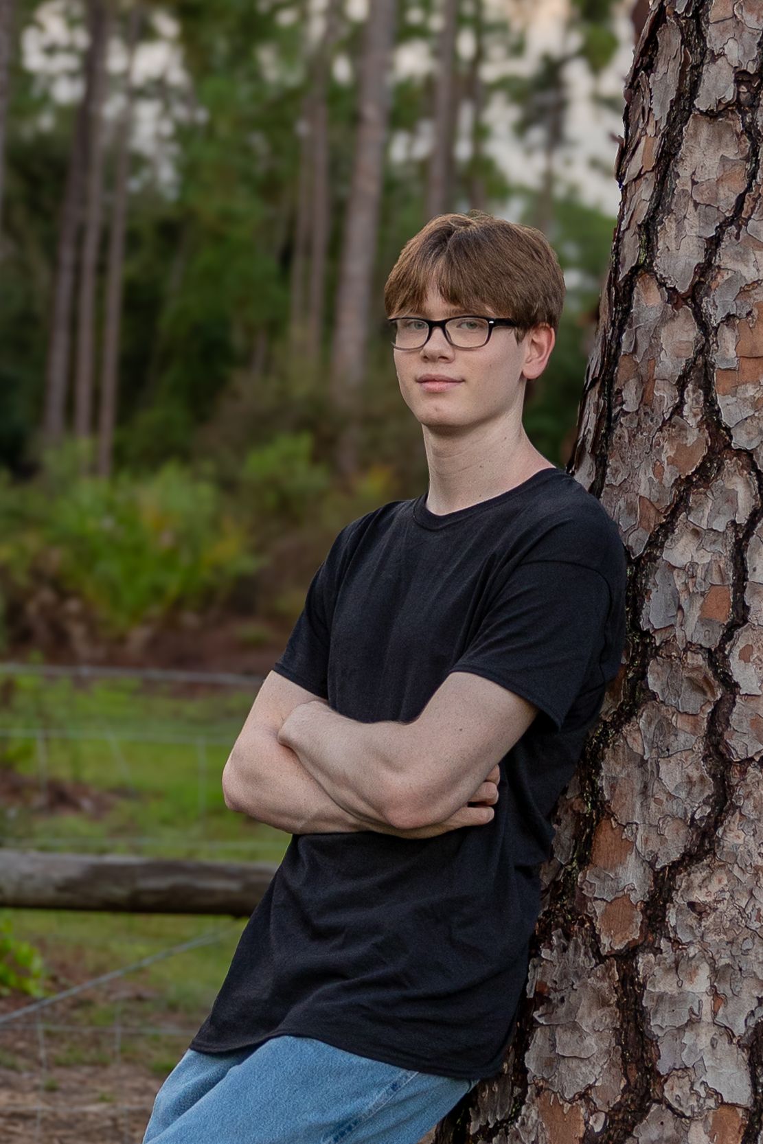 Teen boy with glasses in black t-shirt leaning against pine tree with arms crossed in outdoor setting.
