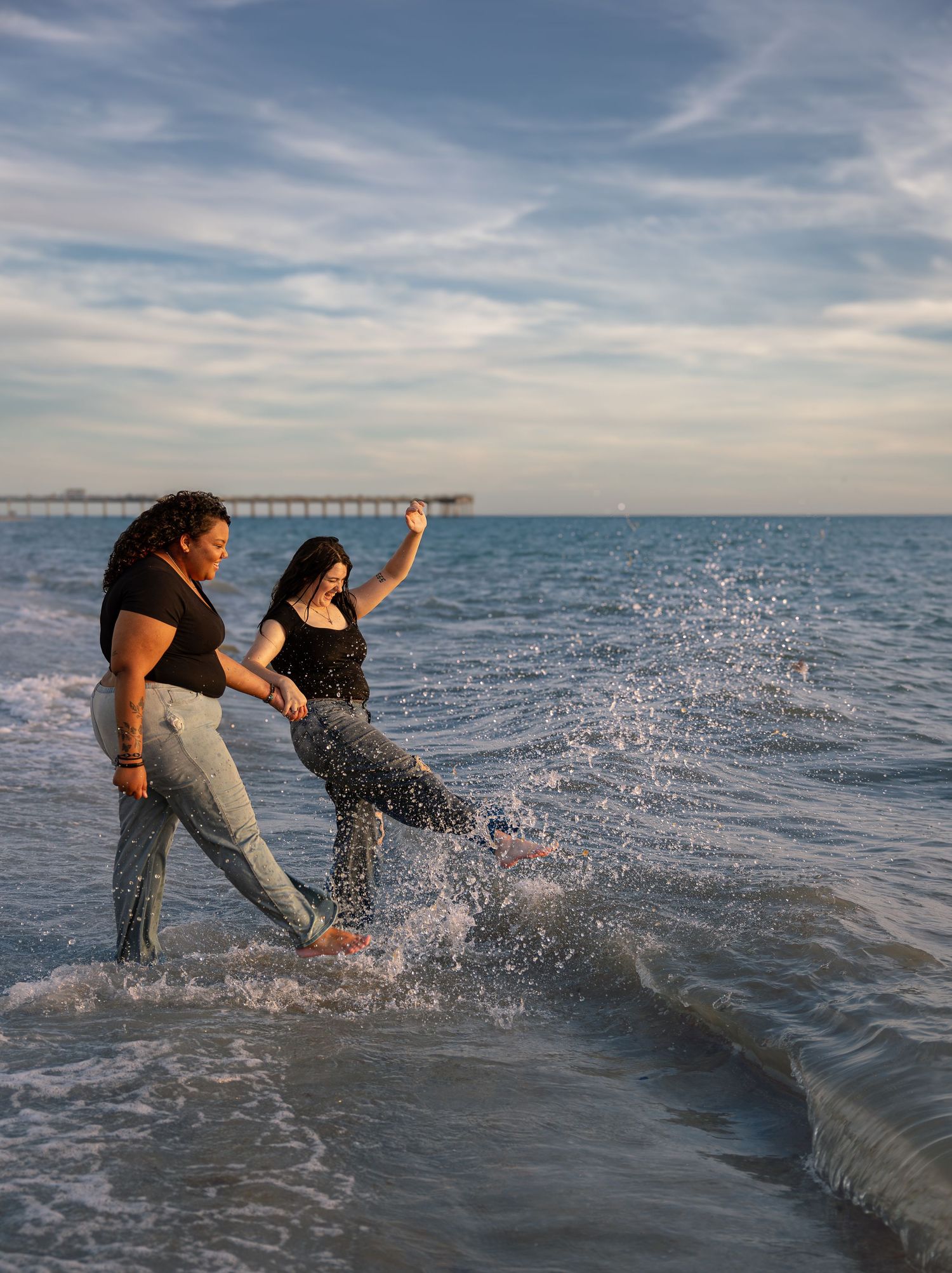 Two women splashing in ocean waves near a pier on a cloudy day.