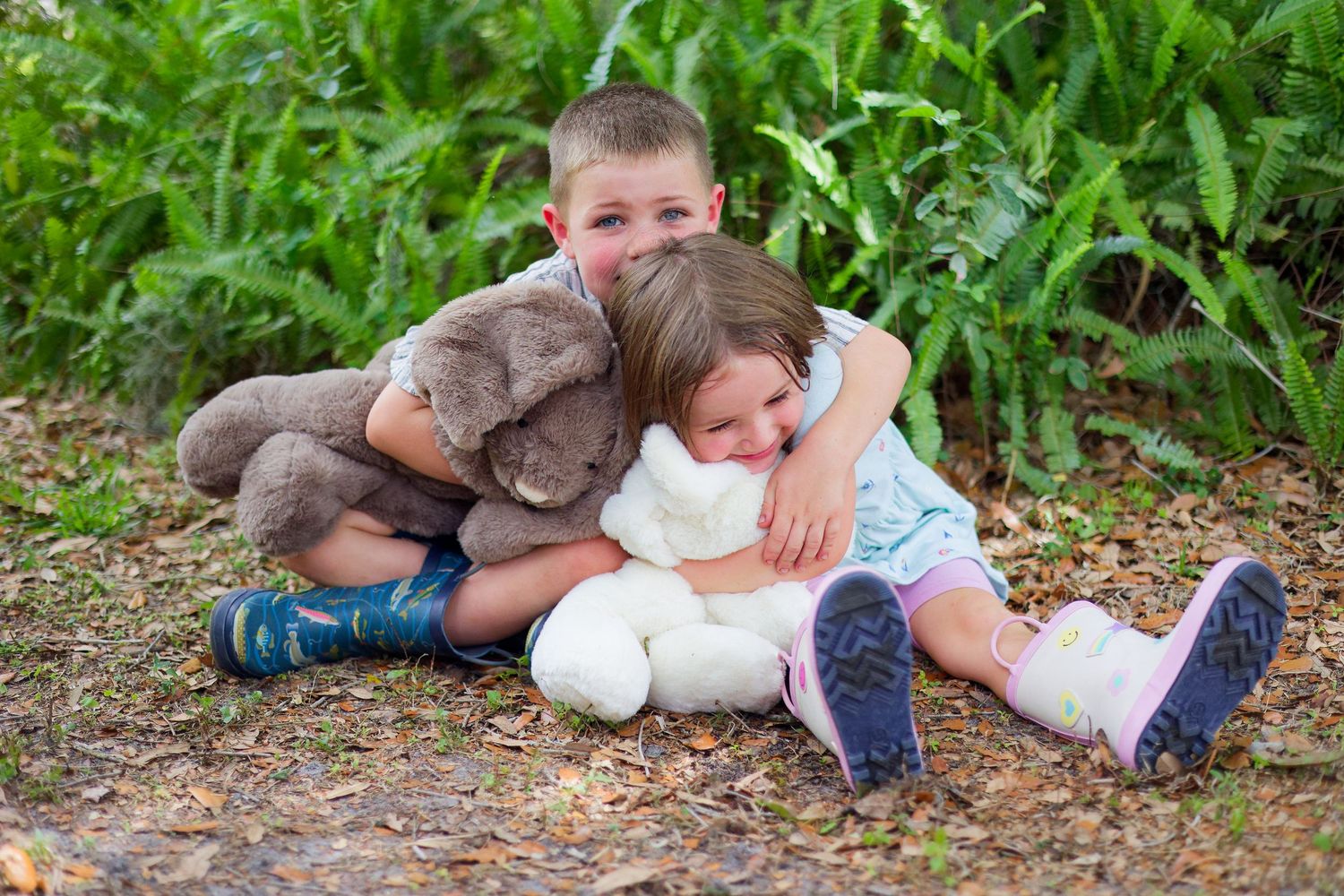 Two children sit on the ground hugging stuffed animal toys surrounded by lush green foliage outdoors.