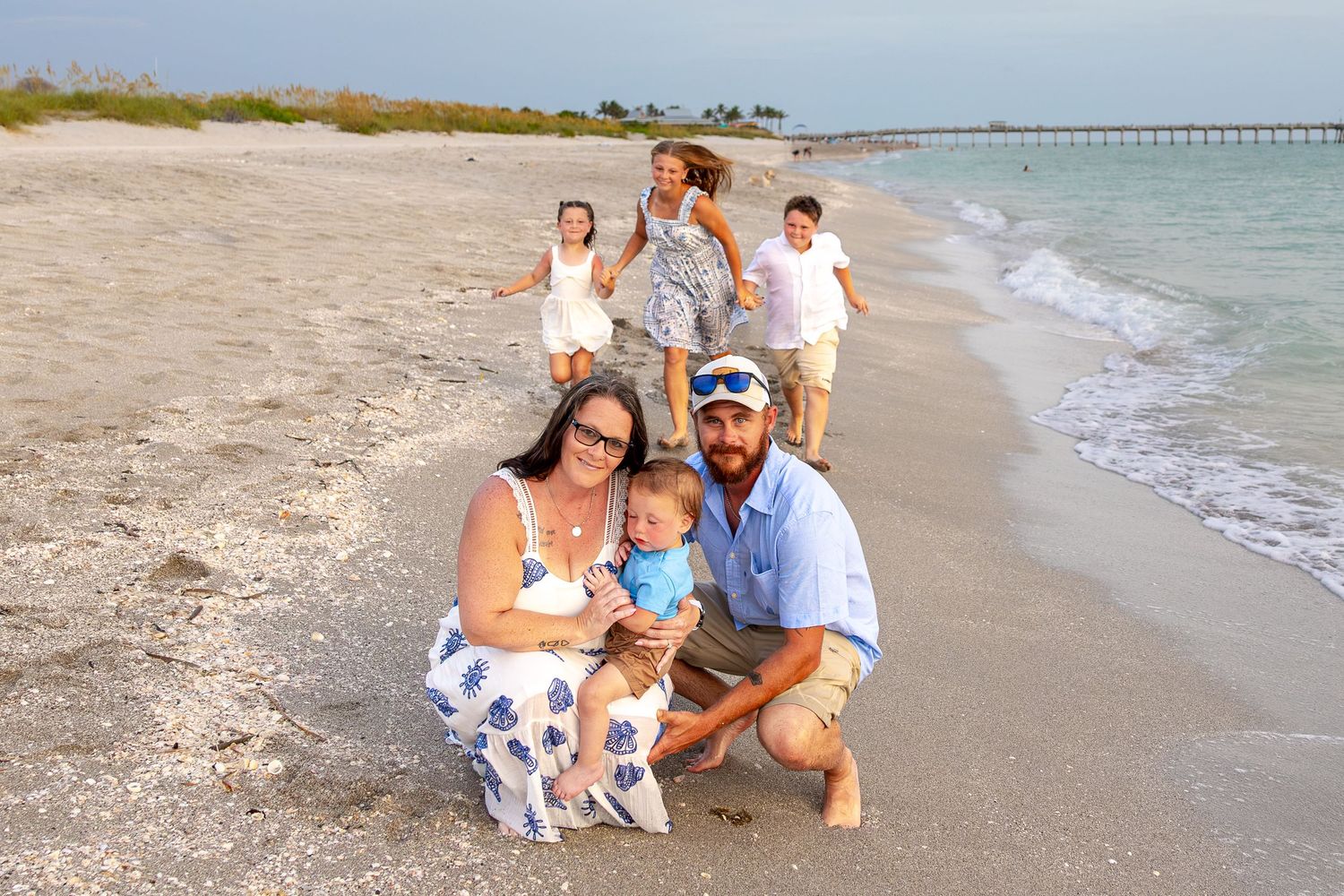 Family posing on a sunny beach with a pier in the background, parents crouching with baby while children play behind them.