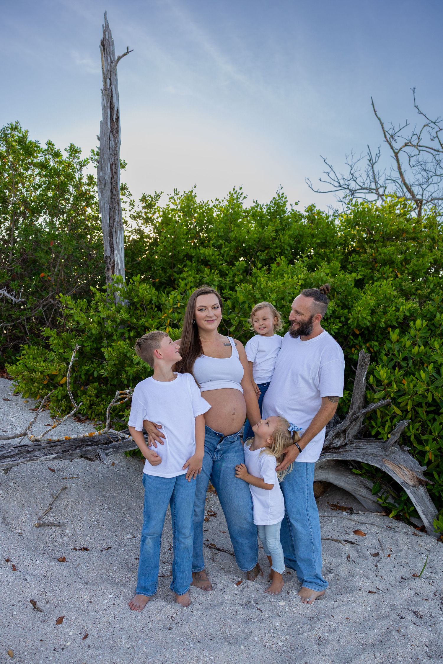 Pregnant woman in white top and jeans poses with family on sandy beach near green bushes.