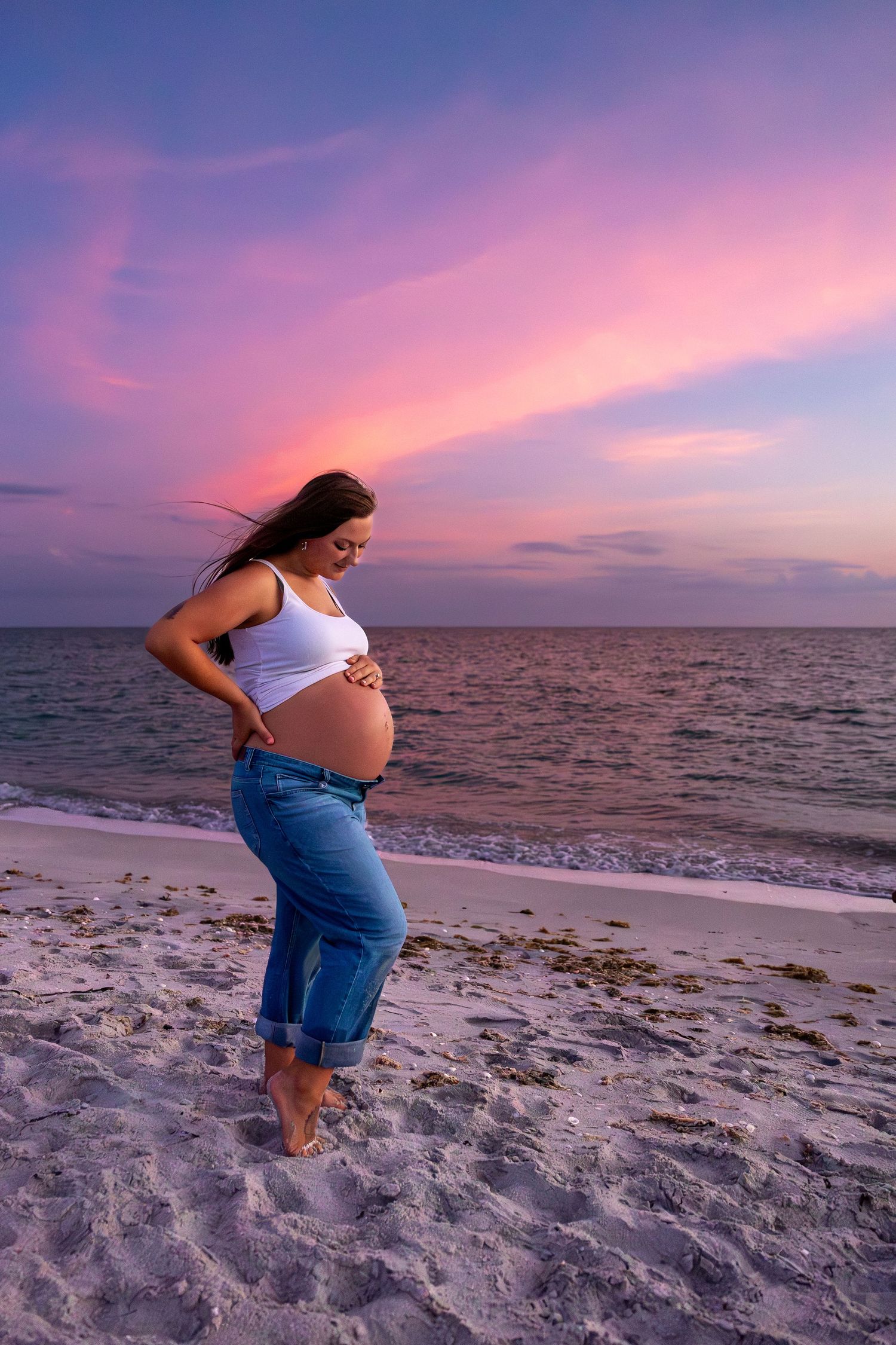 Pregnant woman in white top and jeans cradling her belly on a sandy beach during a vibrant pink and purple sunset.
