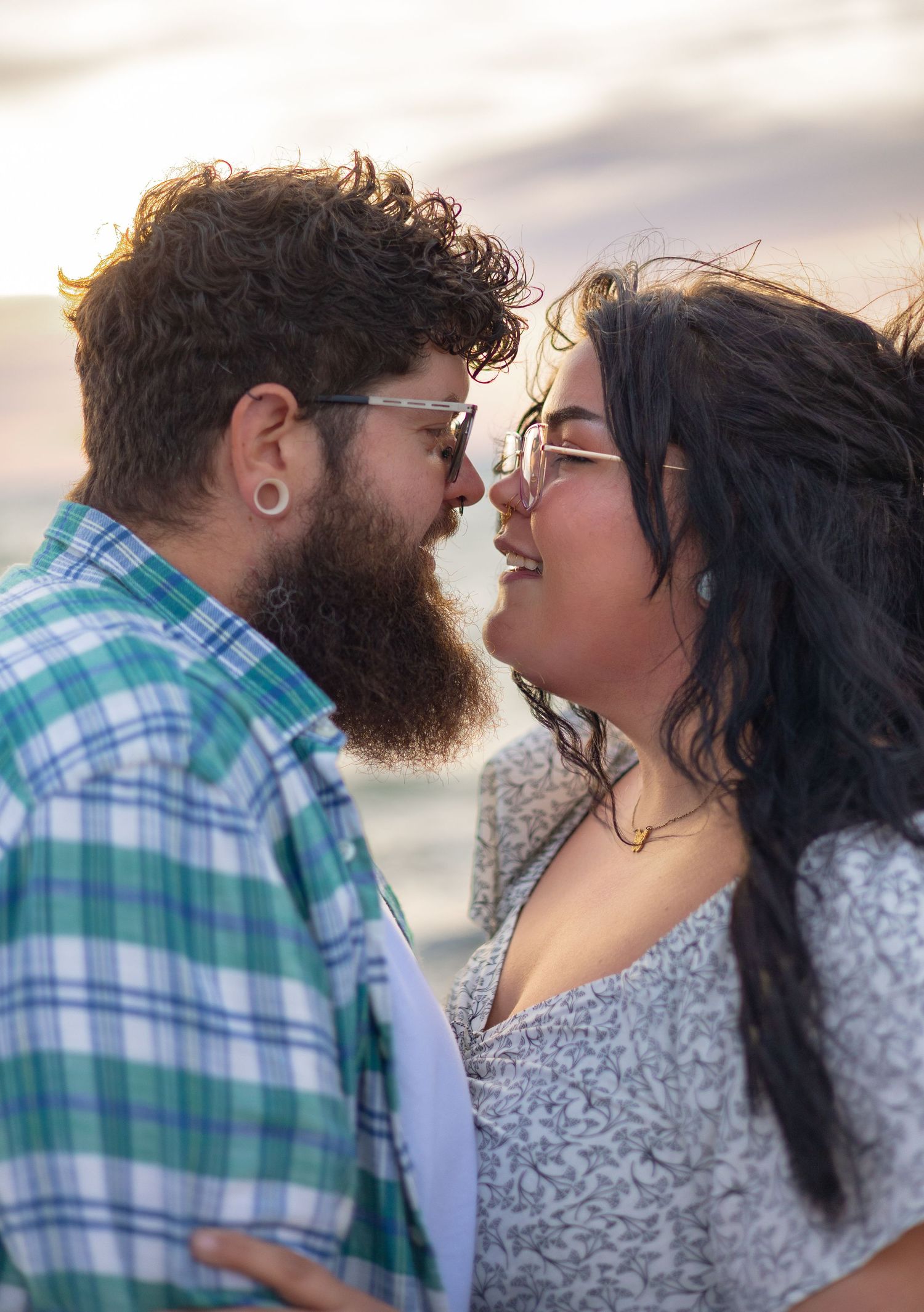 Couple with beards and glasses sharing a romantic moment at the beach during golden hour sunset.