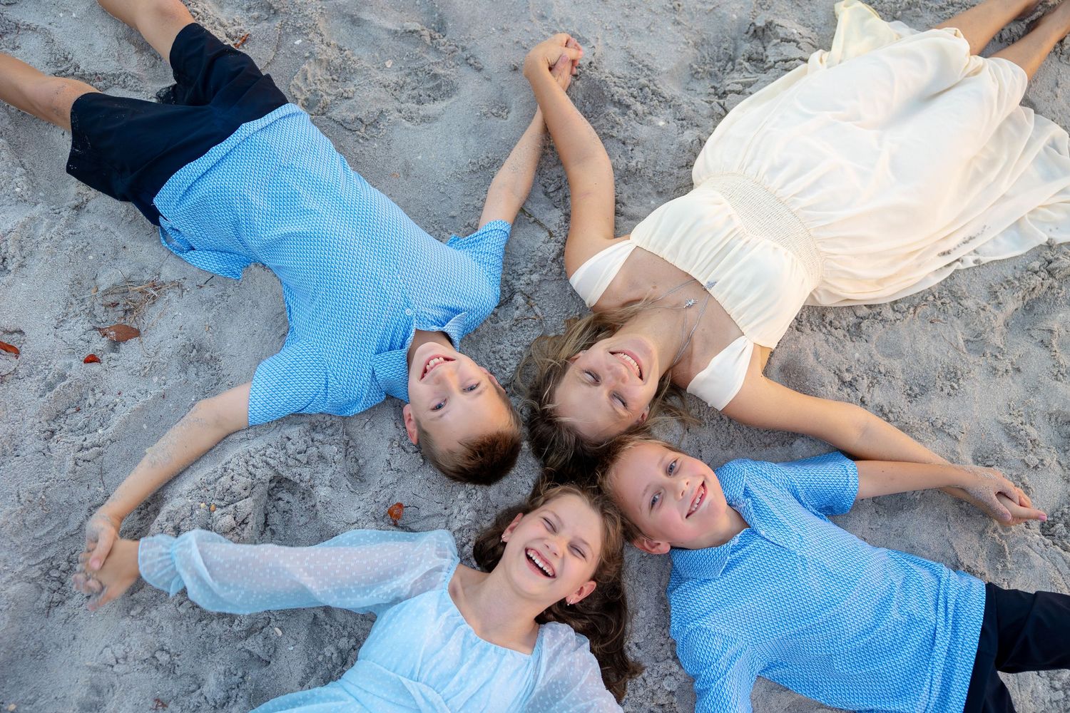 Four children lying on sandy beach in a circle, wearing blue and white outfits, smiling and holding hands.