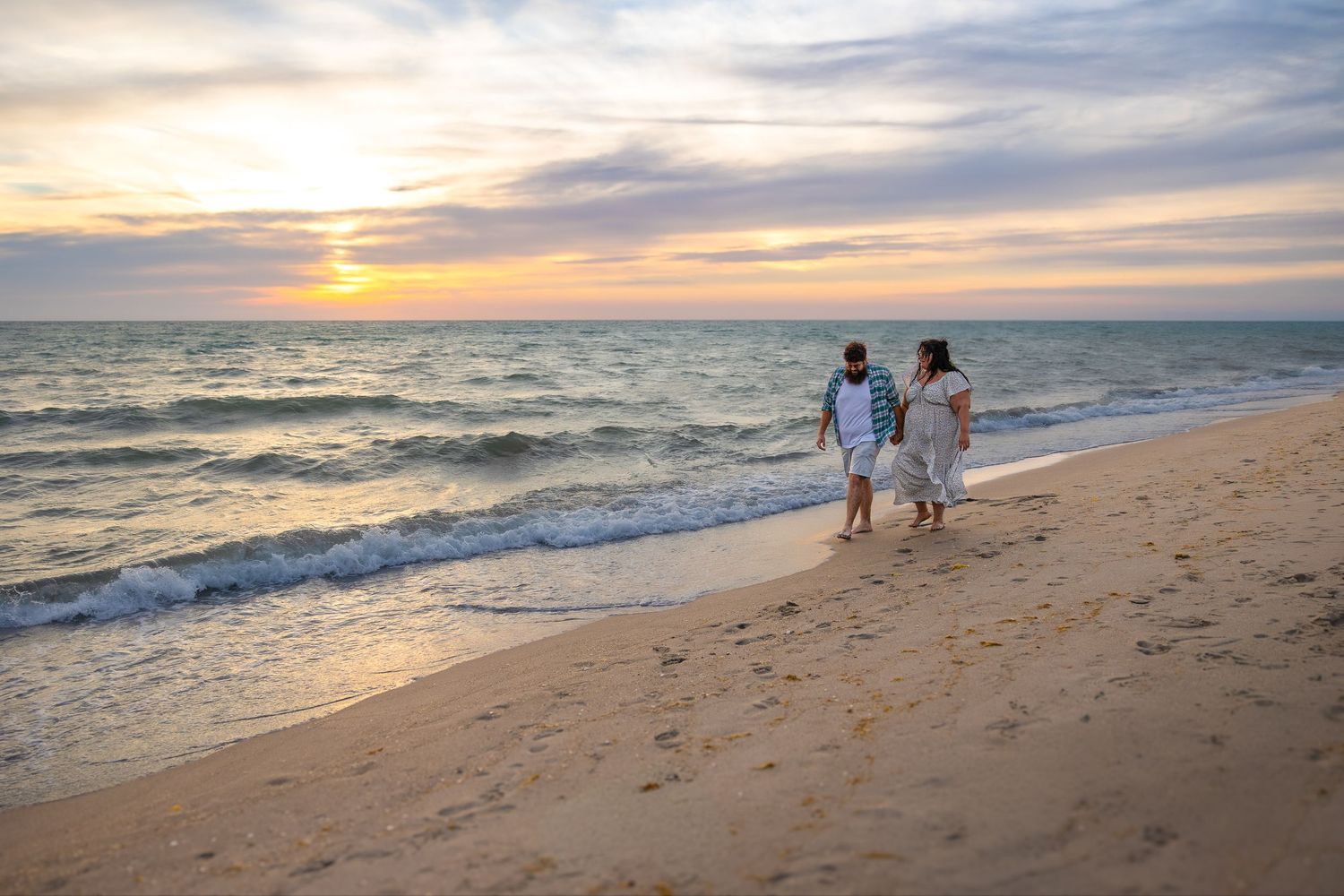 Couple walking hand in hand along a sandy beach shore during a colorful sunset evening.
