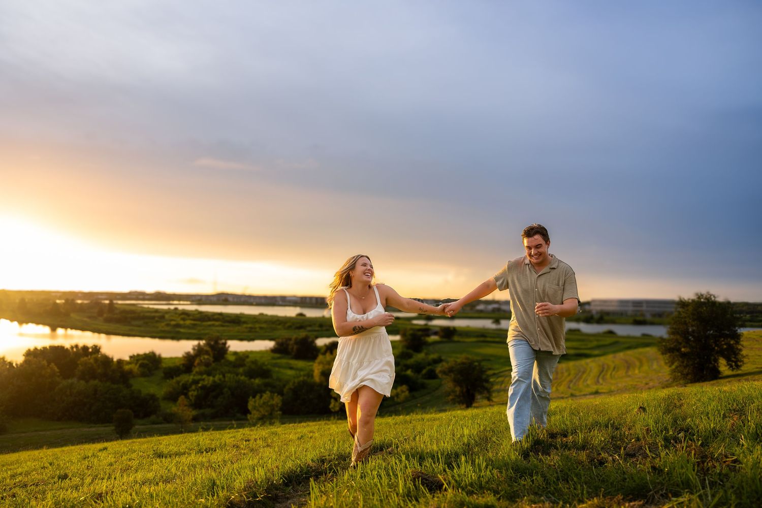 A joyful couple holds hands running through a lush green meadow at golden sunset near a serene lake.