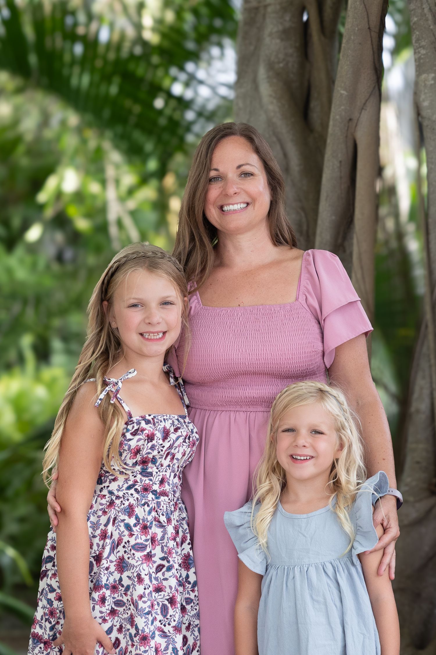 A smiling woman in a pink dress poses outdoors with two blonde girls in floral dresses amid lush tropical greenery.