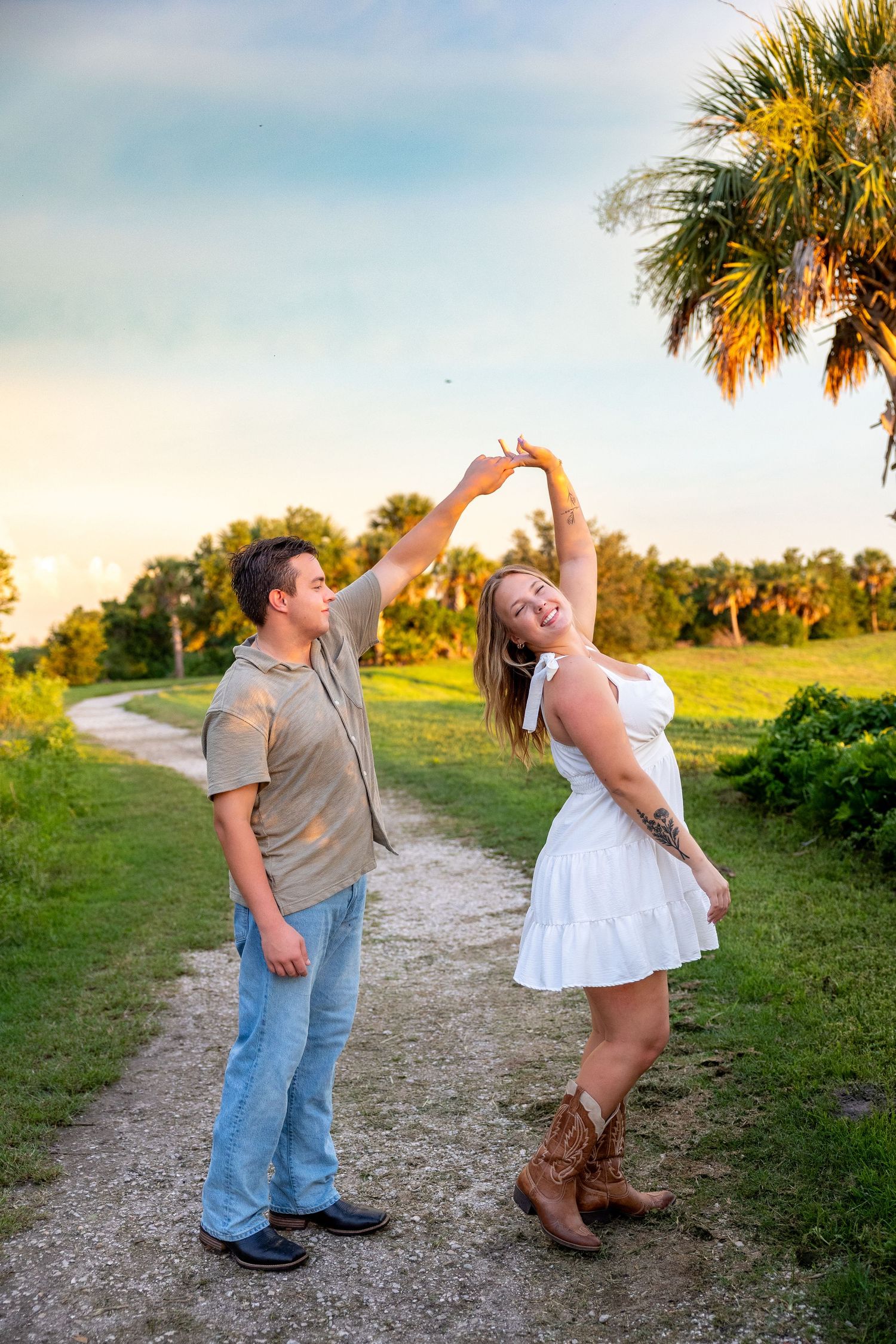 A couple dances on a scenic path at sunset, man spinning woman in white dress and cowboy boots near palm trees.