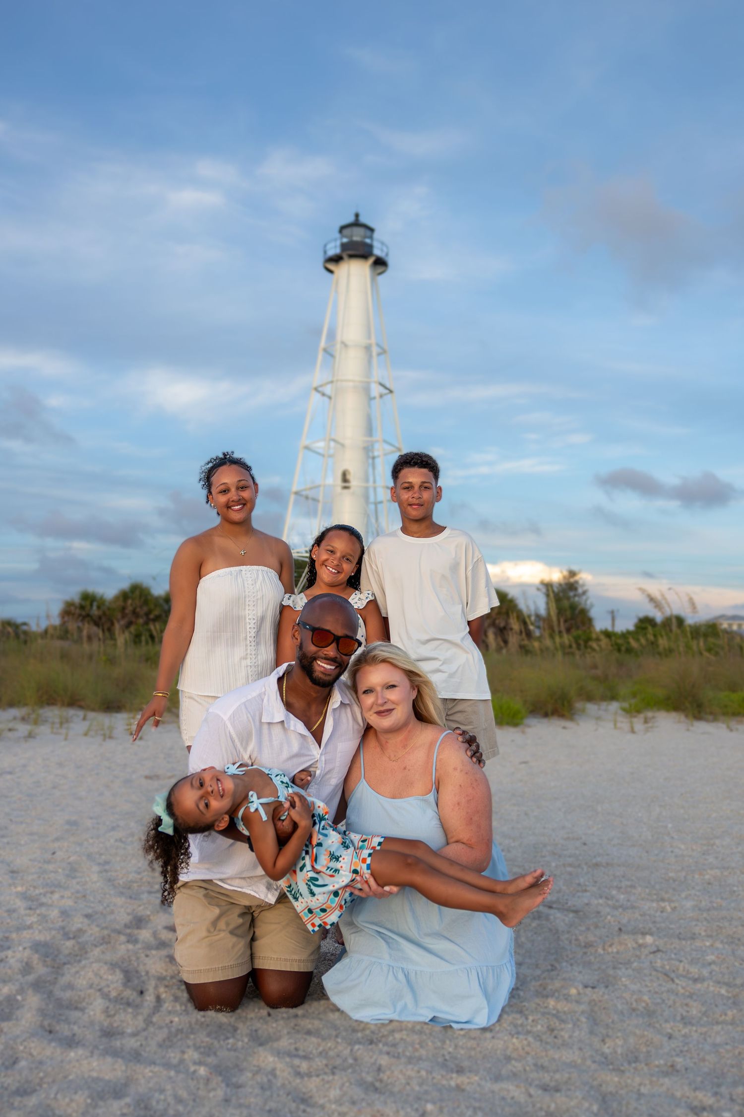 Happy multiracial family posing together on a sandy beach with a white lighthouse backdrop at sunset.