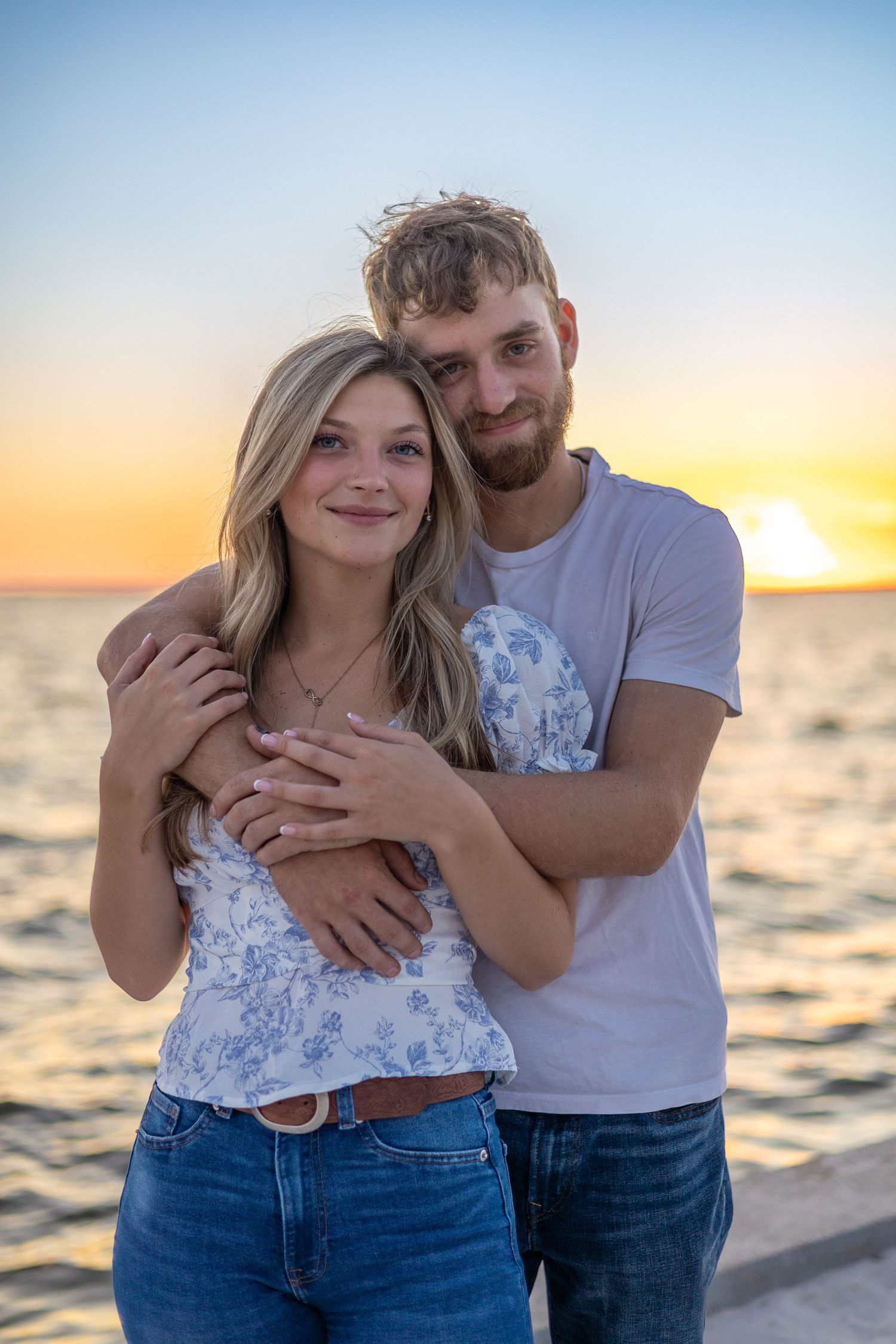 Bearded man embracing blonde woman from behind at oceanside sunset.