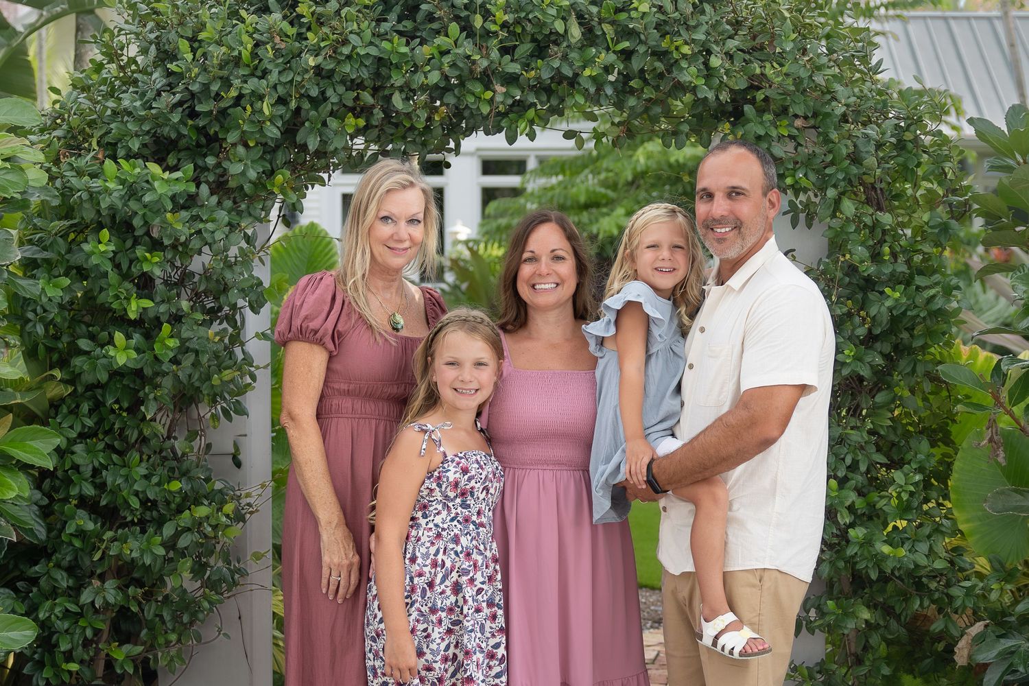 Family of five smiling under a lush green garden arch, with two girls and a boy, dressed in pink and mauve tones.