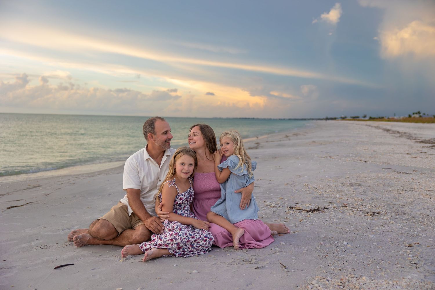 Family of four sitting on a sandy beach at sunset, two young girls smiling with parents on a calm shoreline.