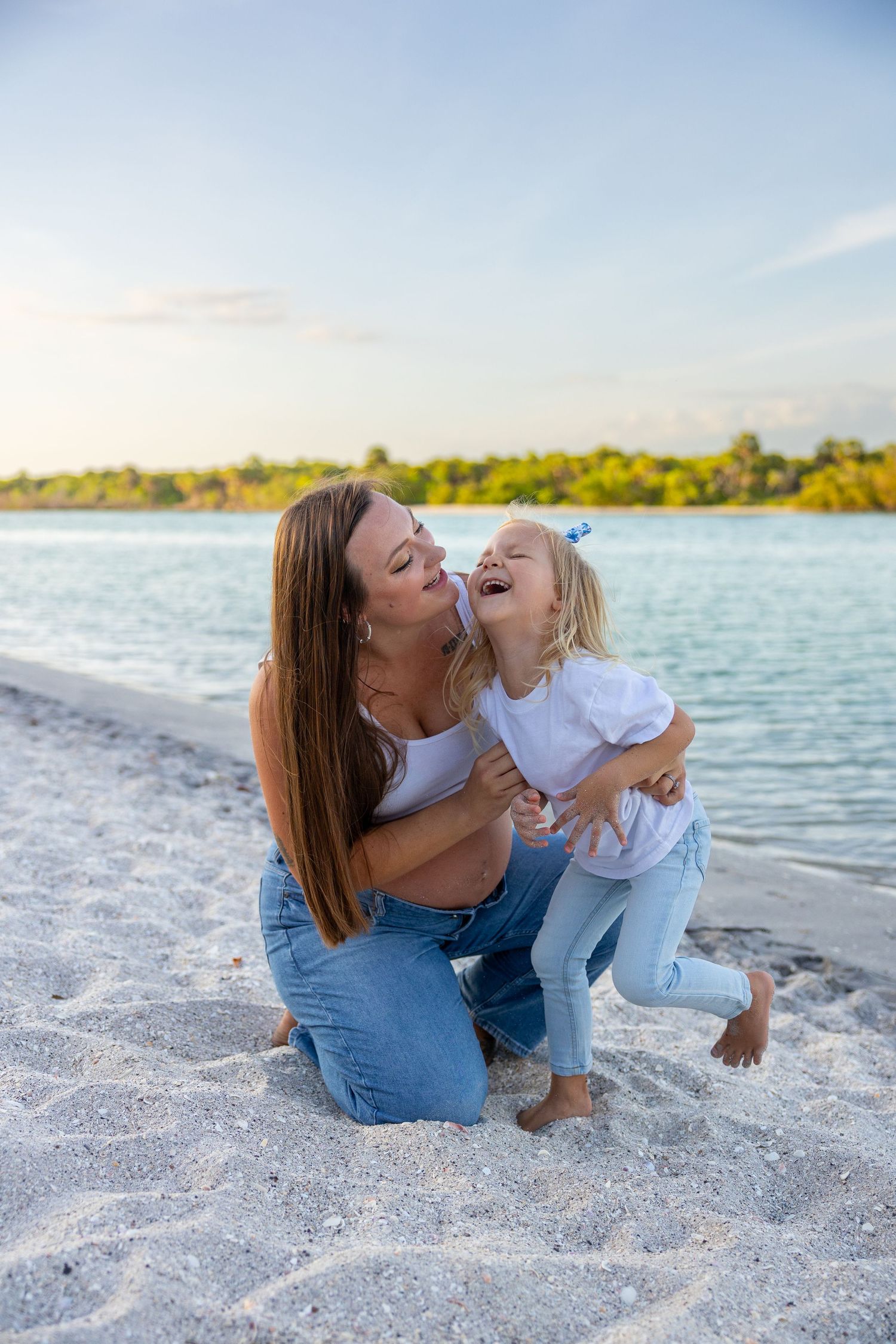 Pregnant mother and toddler daughter laughing together on sandy beach near turquoise water at sunset.