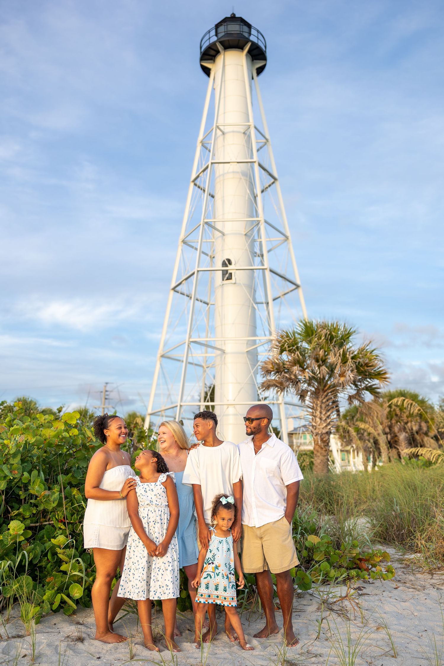 Family of six poses barefoot before a tall white lighthouse surrounded by palm trees on a sunny beach day.