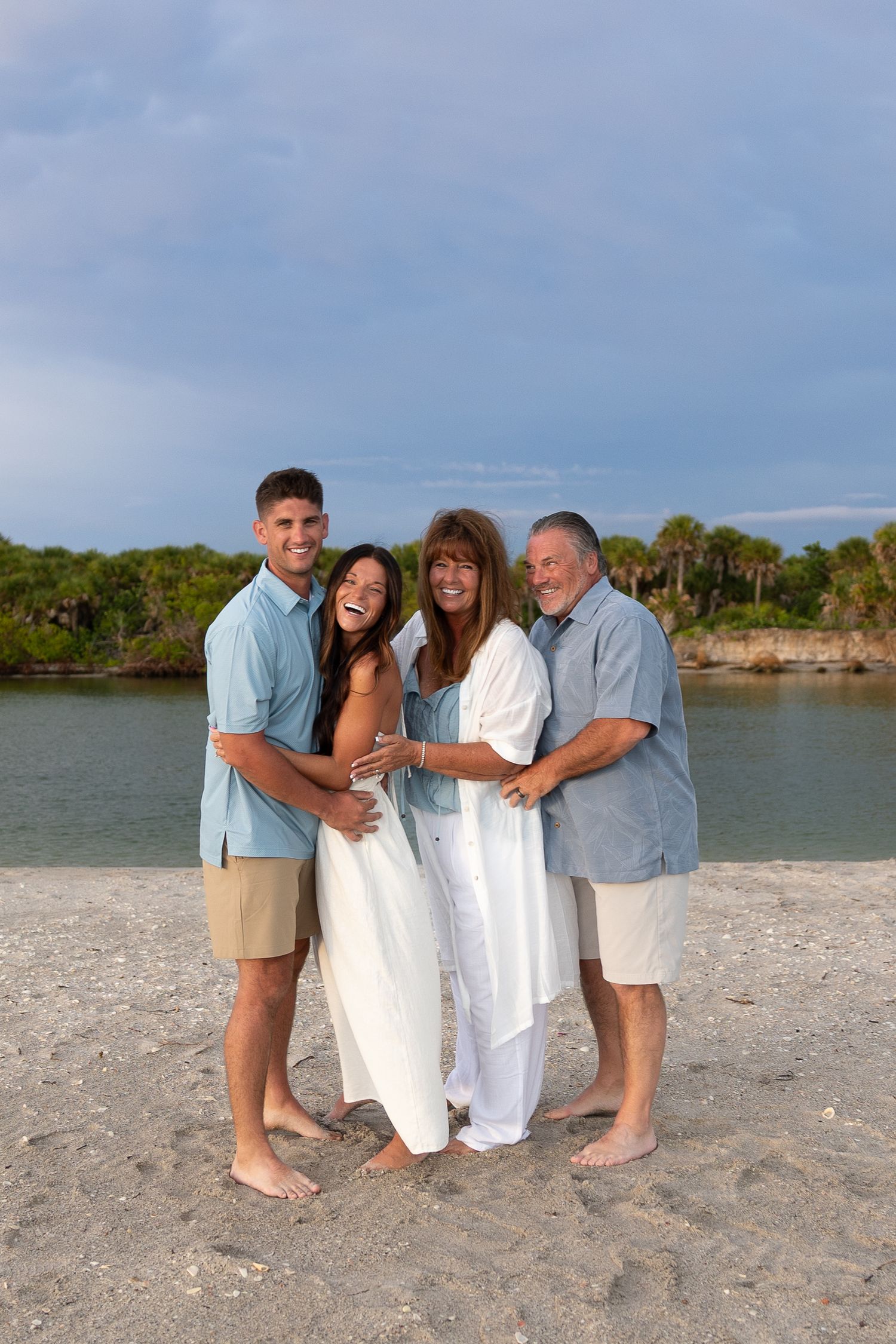 Happy family of four posing together on a sandy beach with calm water and palm trees in the background.