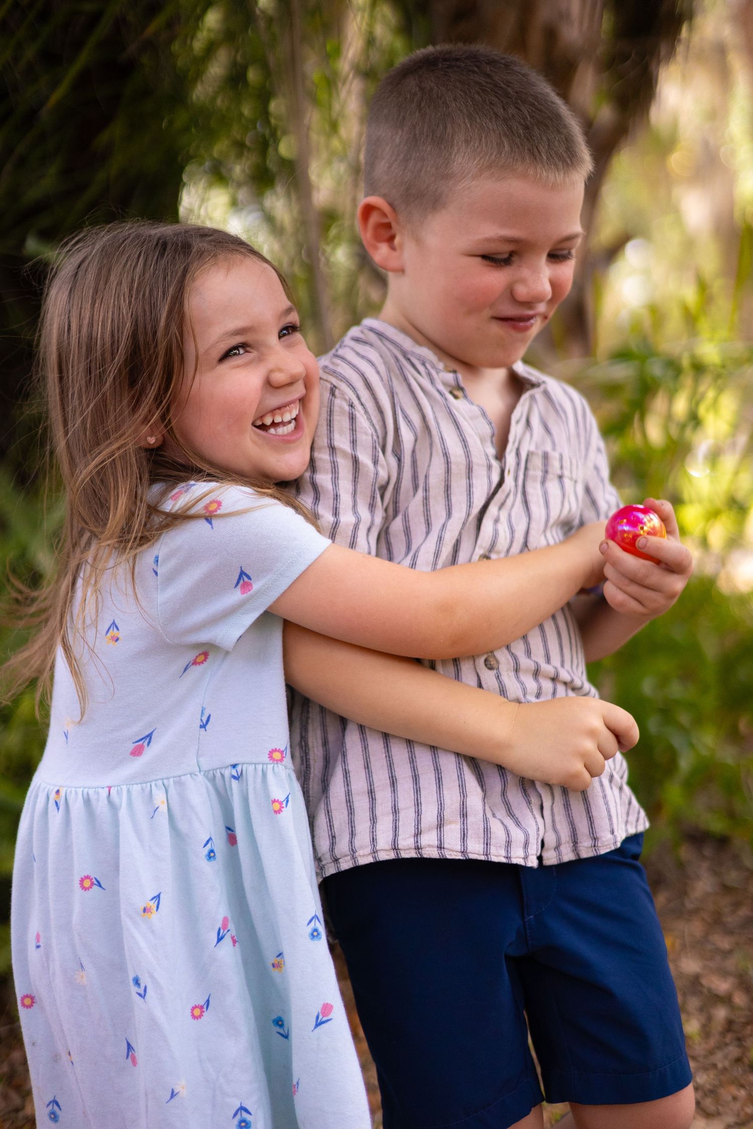 Two young children hugging and laughing outdoors, girl in blue dress embracing boy holding pink candy.