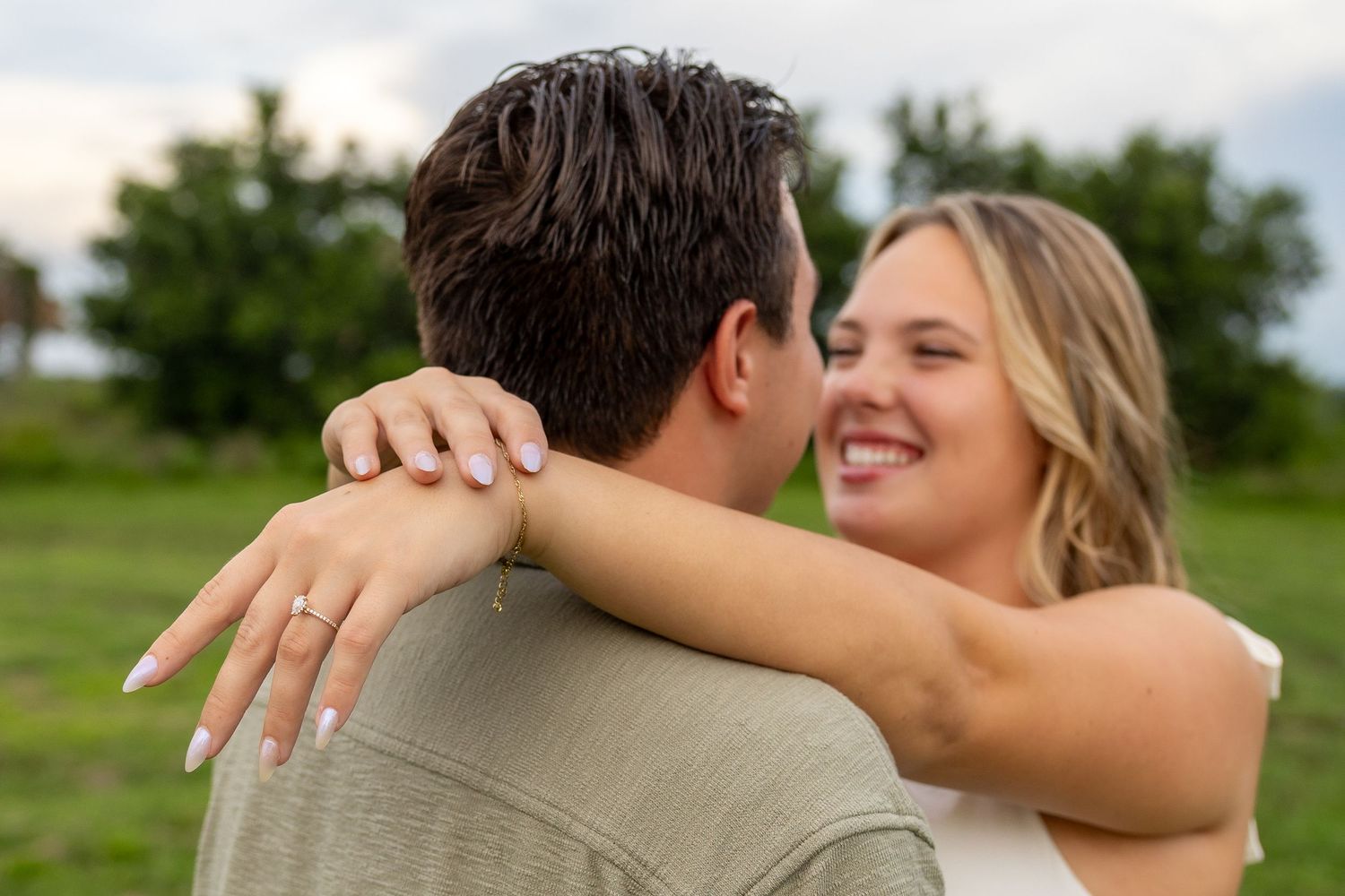 Couple embracing outdoors, woman smiling with engagement ring visible on hand around man's neck, green field background.