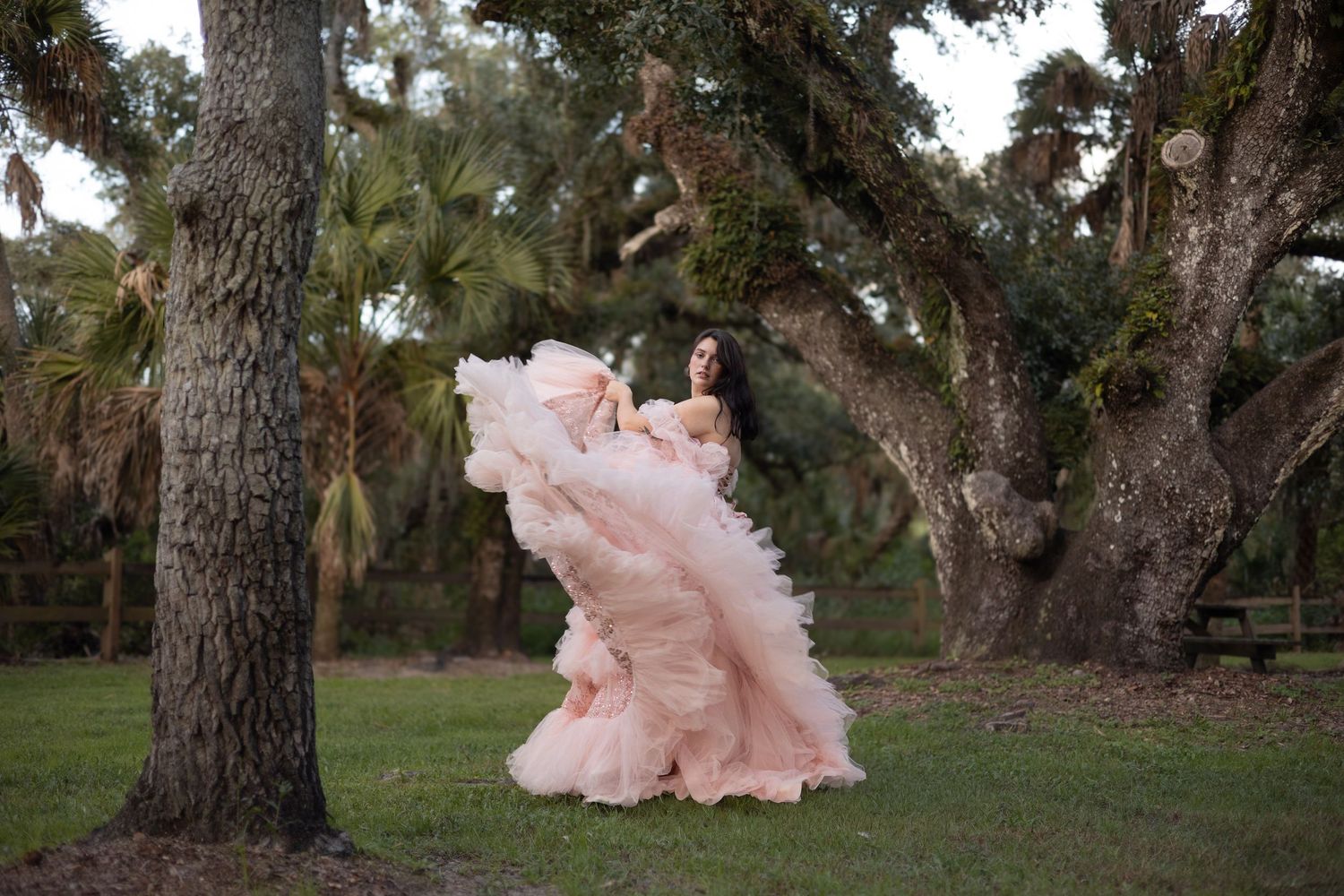 Woman in flowing pink ball gown poses dramatically among large oak trees in a lush green outdoor setting.