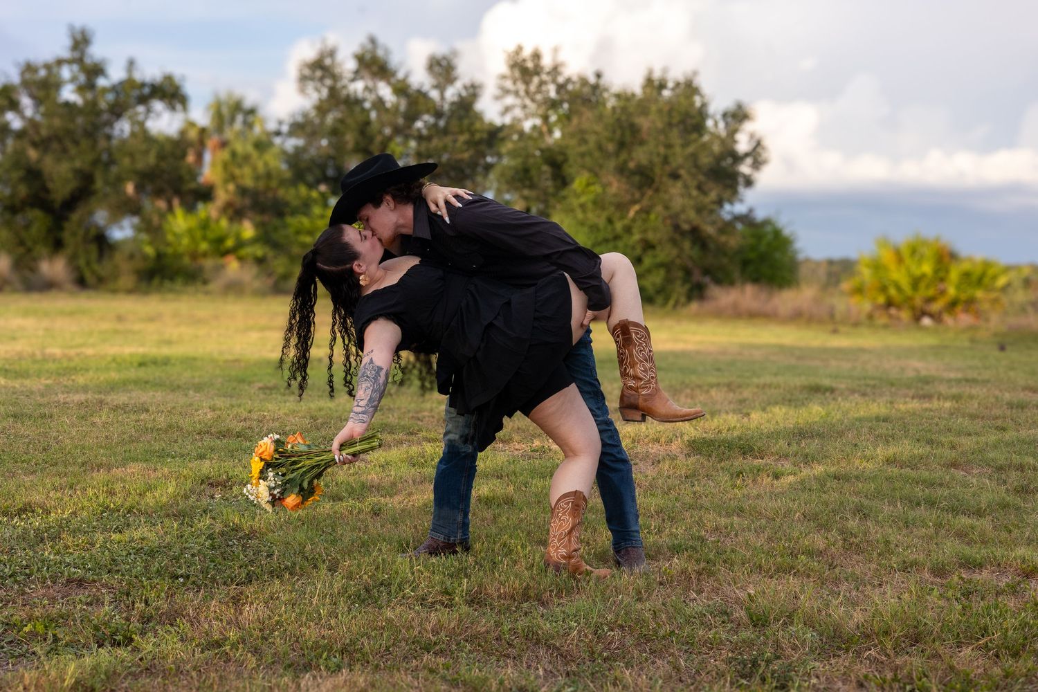 Couple sharing a romantic dip kiss in a green field, woman holding sunflowers, wearing cowboy boots and black outfits.