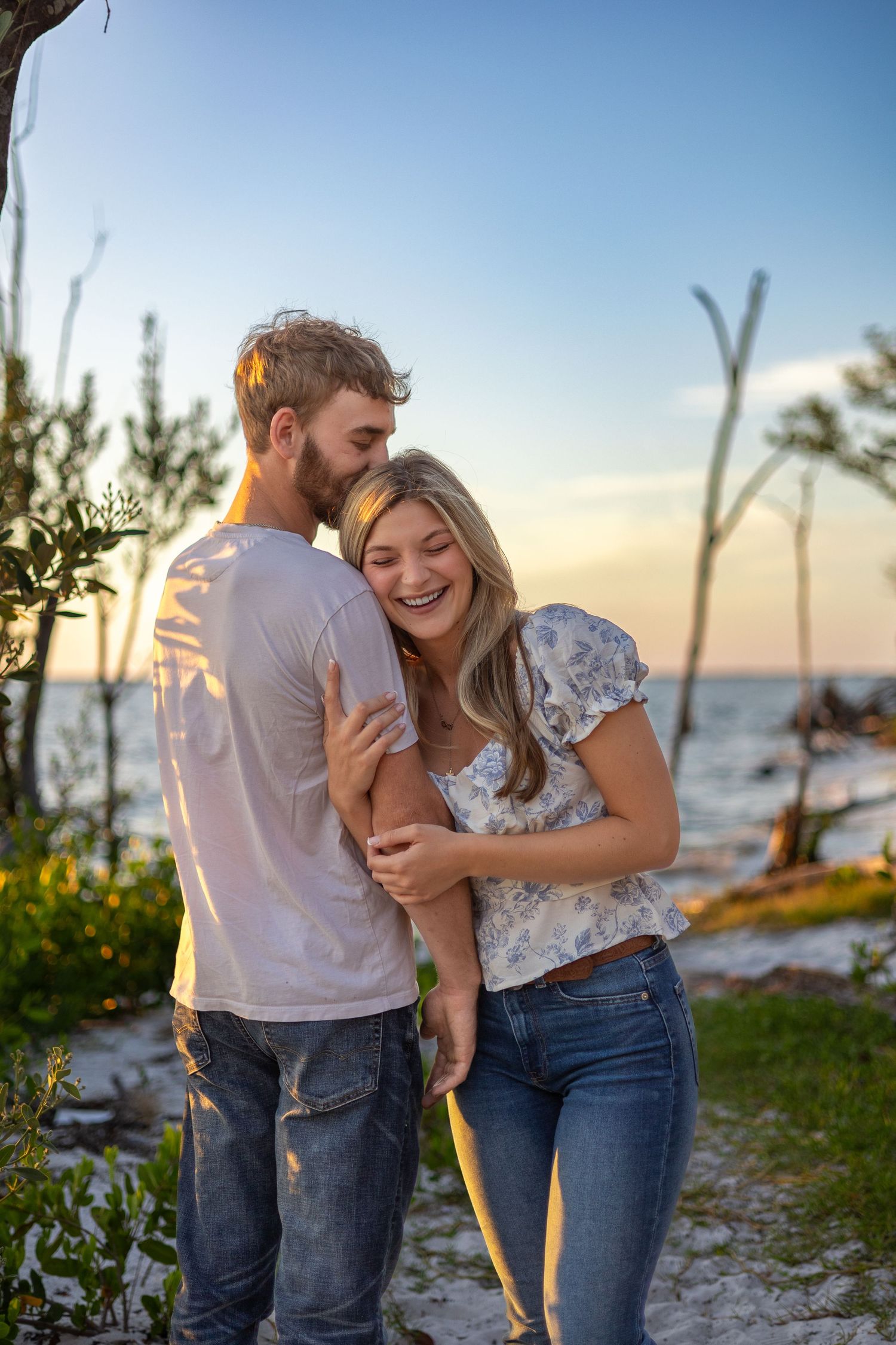 Laughing couple shares a tender moment on a beach at sunset with sparkling ocean in background.