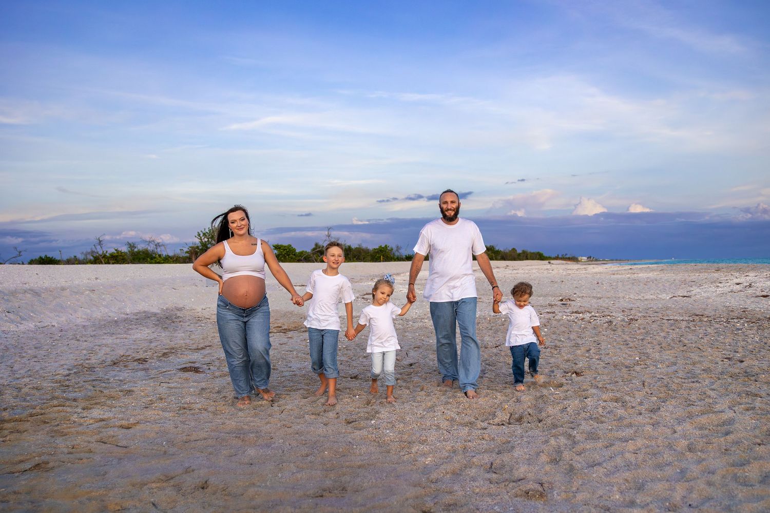 Pregnant woman and bearded man walk on beach holding hands with three young children under blue sky.