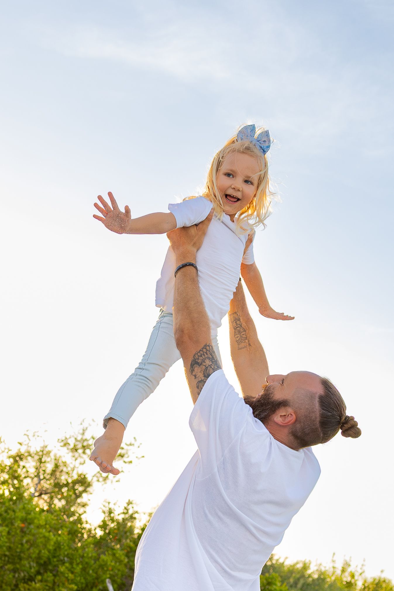 A bearded father joyfully lifts his laughing blonde daughter wearing a bow into the sky outdoors.