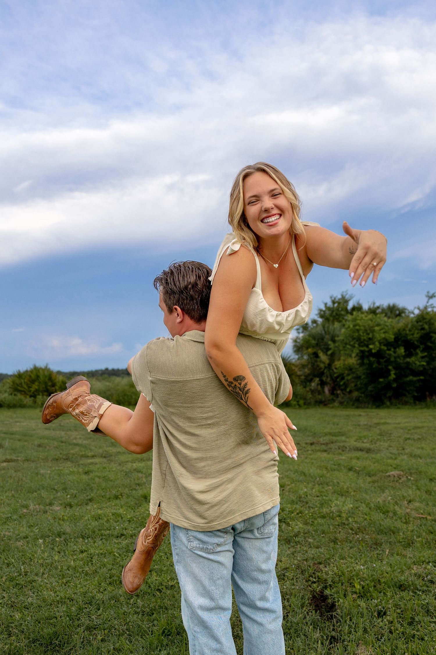 A man lifts a smiling blonde woman in a green field under a cloudy sky, both wearing casual summer outfits.