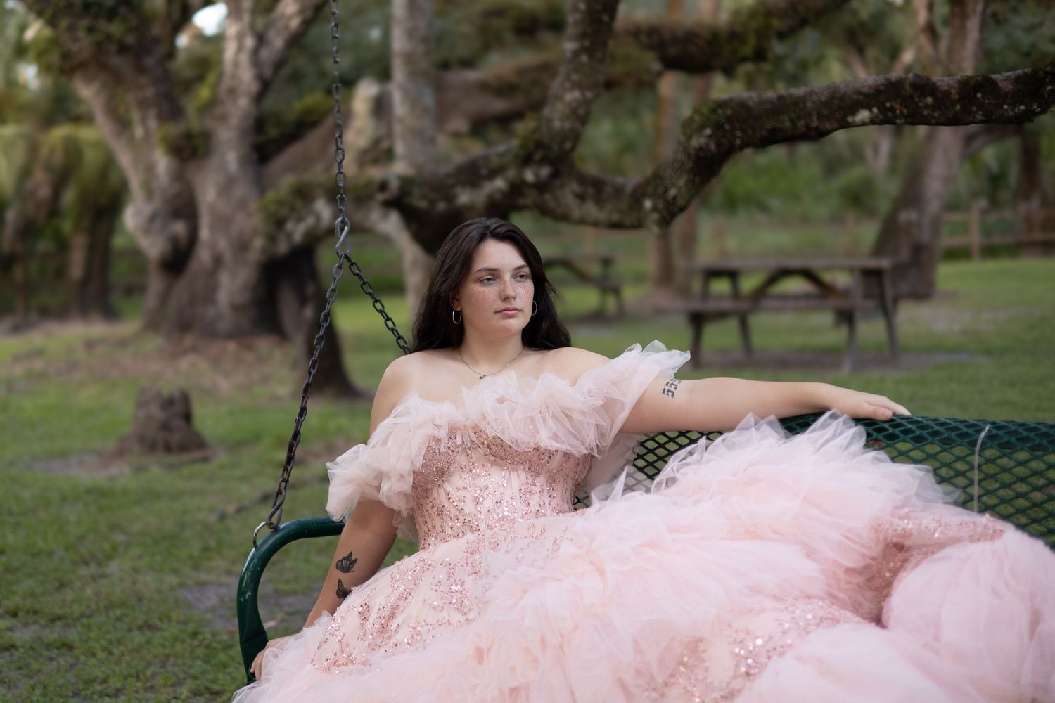 Young woman in a voluminous blush pink tulle gown sitting on a garden swing surrounded by lush green trees.
