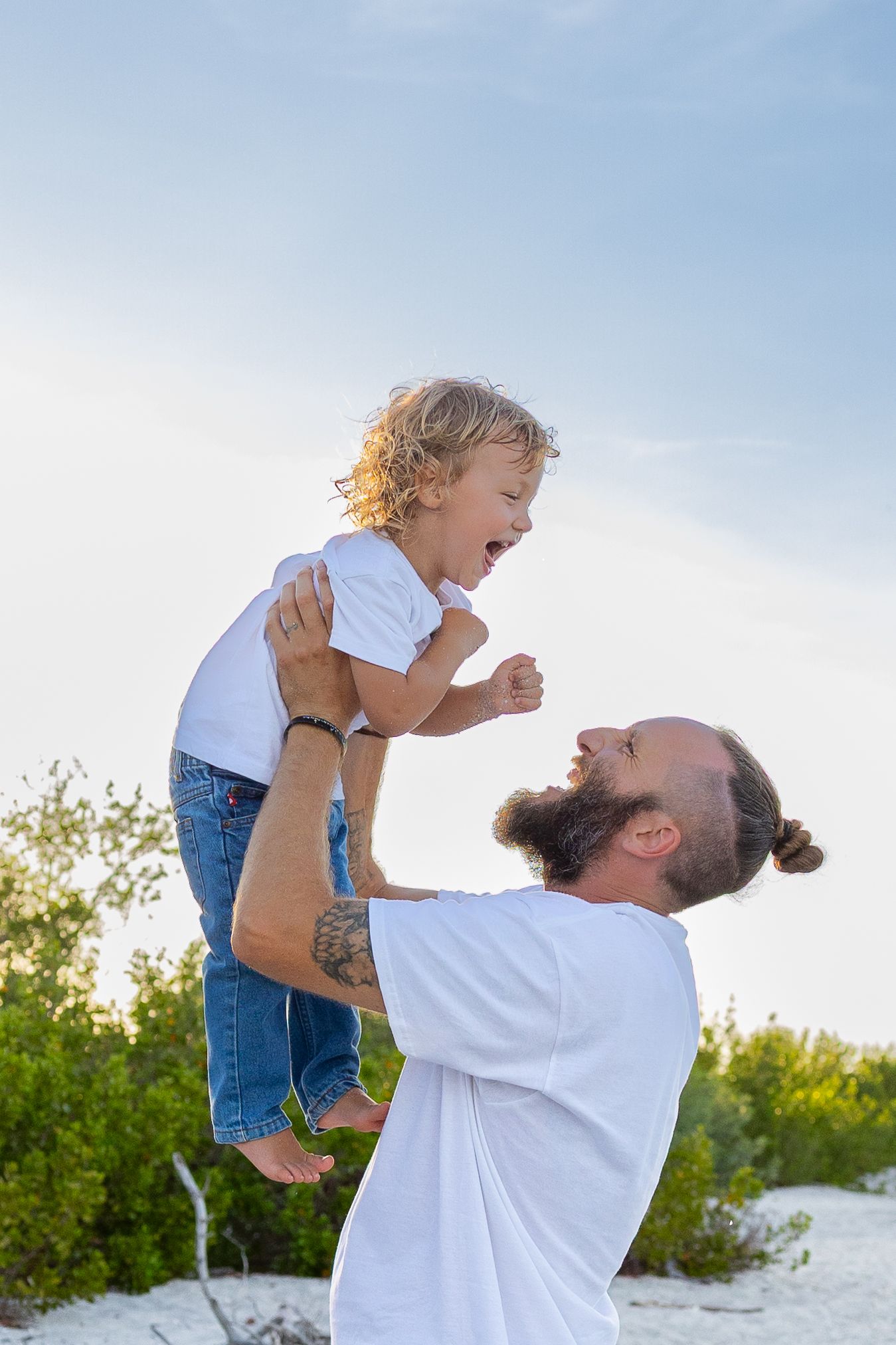 Bearded father lifting laughing toddler boy in white shirt outdoors against blue sky near green bushes.