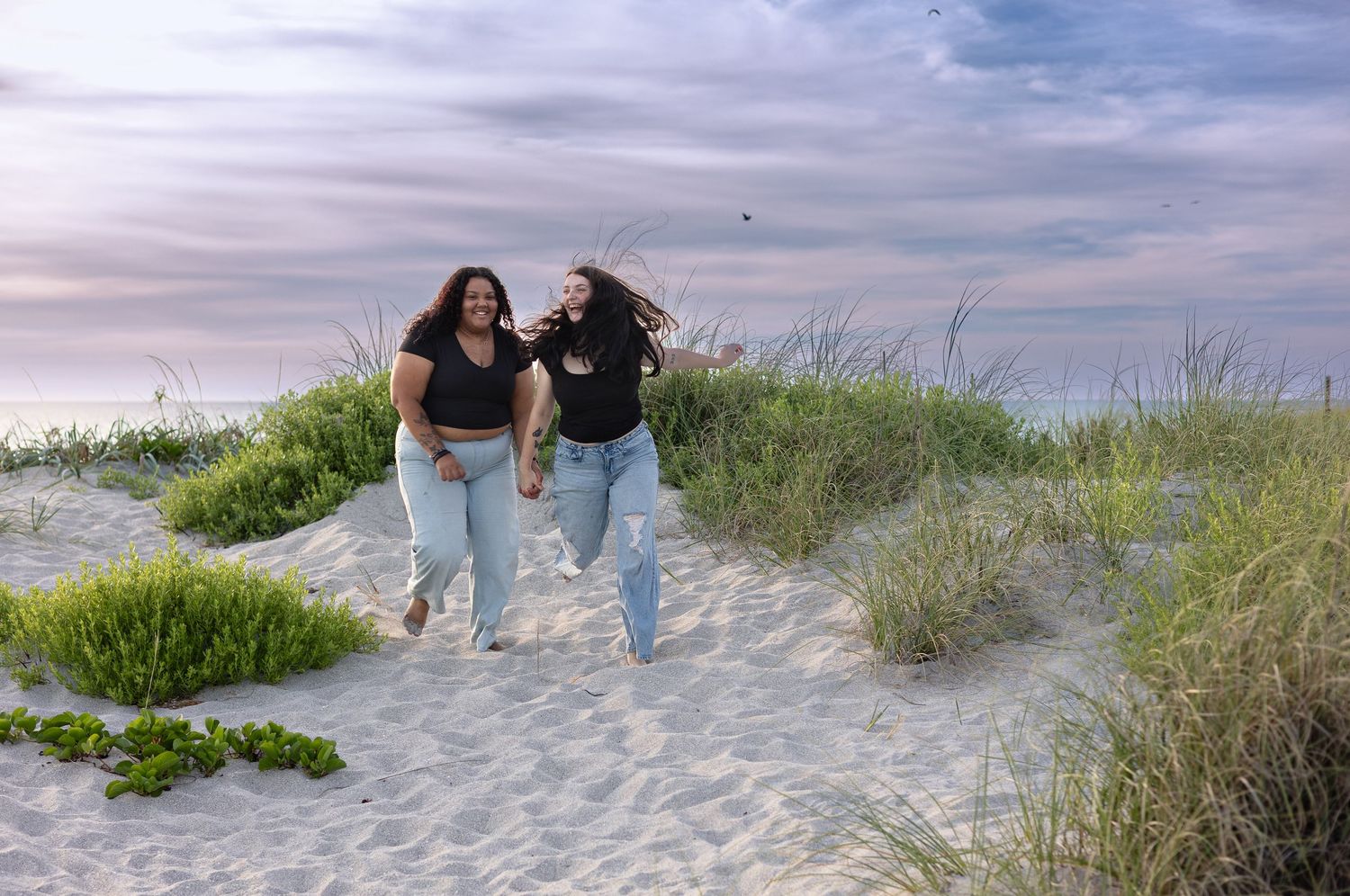Two happy women running together through grassy sand dunes at sunset beach.