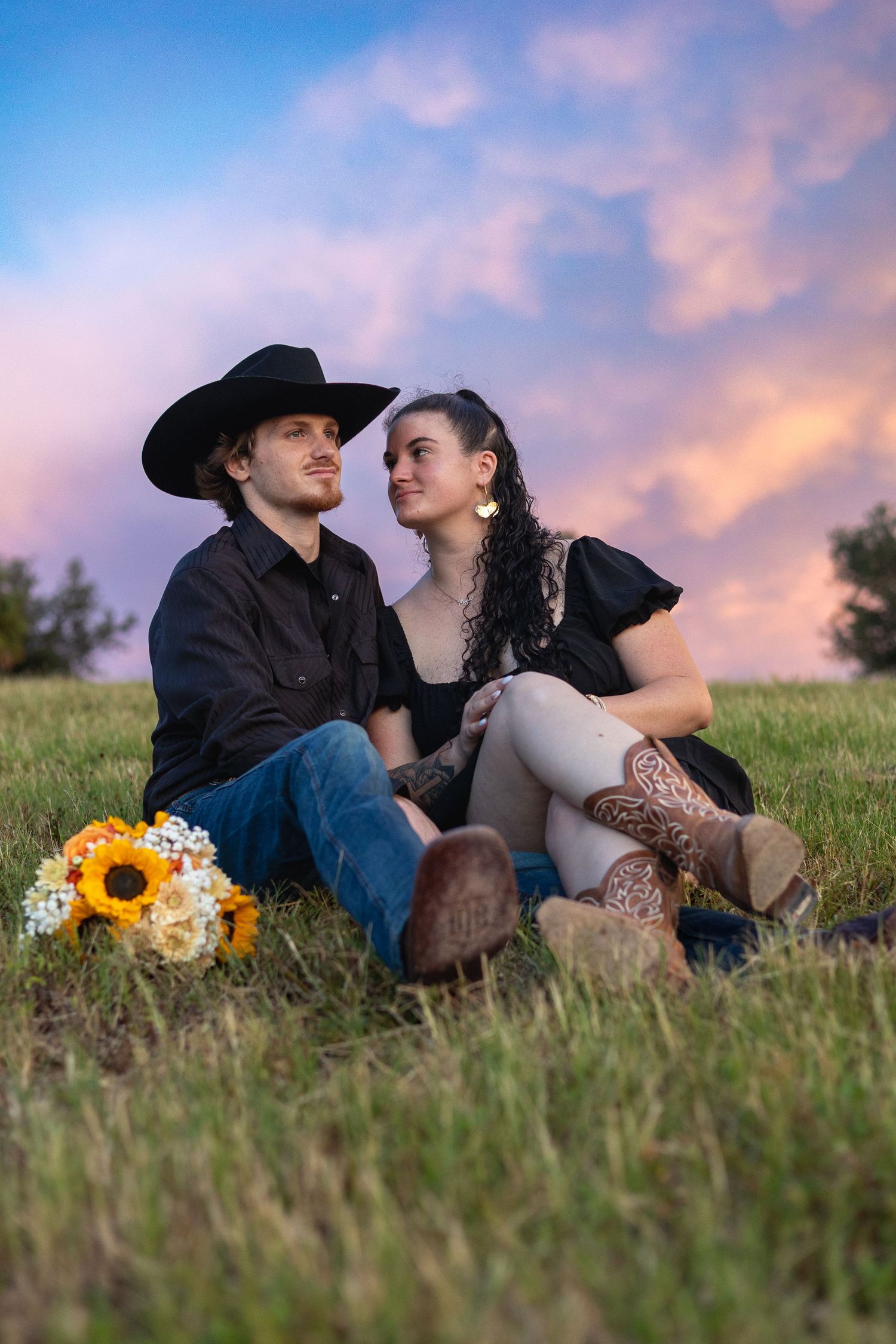 Couple in western attire sitting on grass at sunset, man in cowboy hat, woman in boots, with sunflower bouquet nearby.