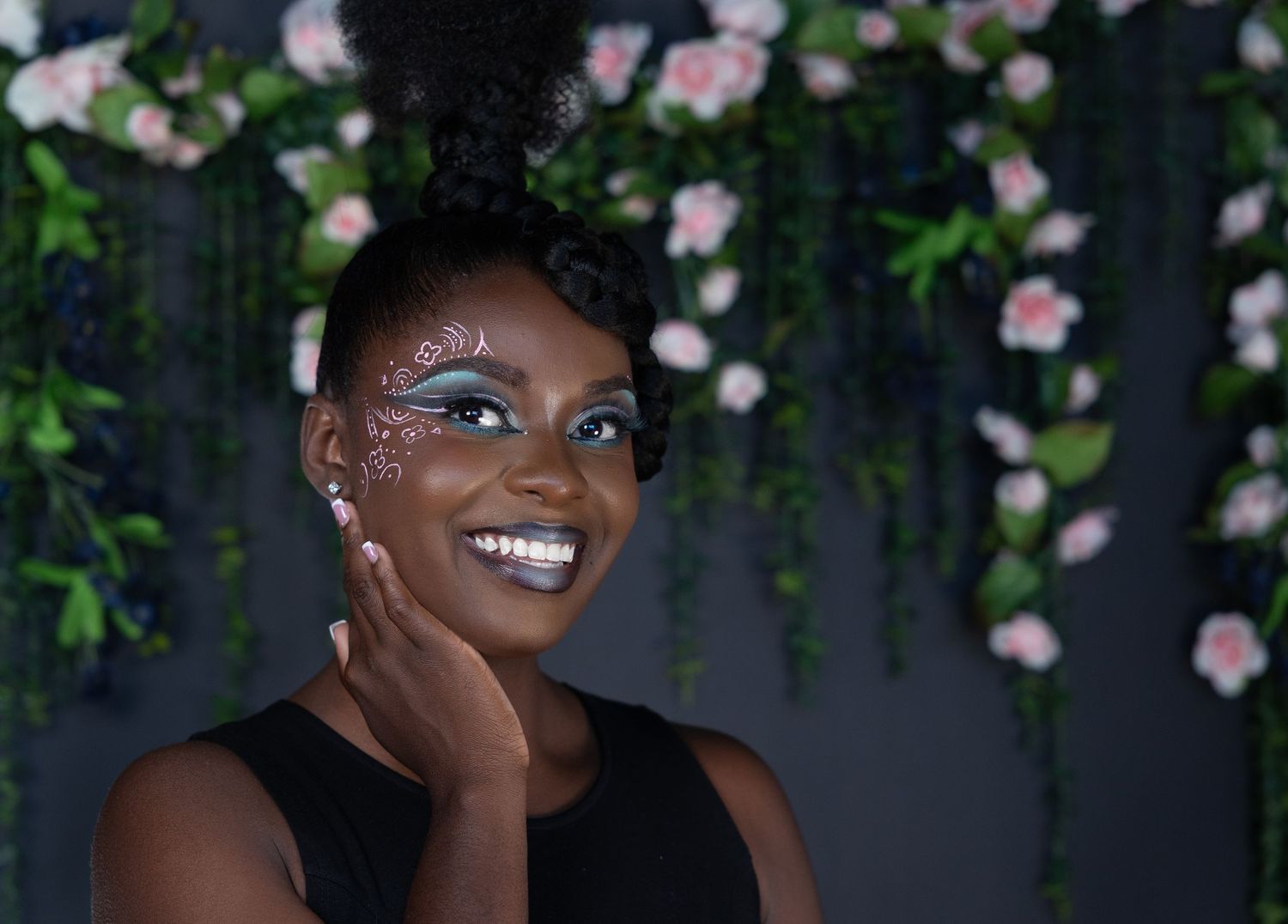 Woman with artistic face gems and teal eye makeup smiles against a floral backdrop with pink roses.