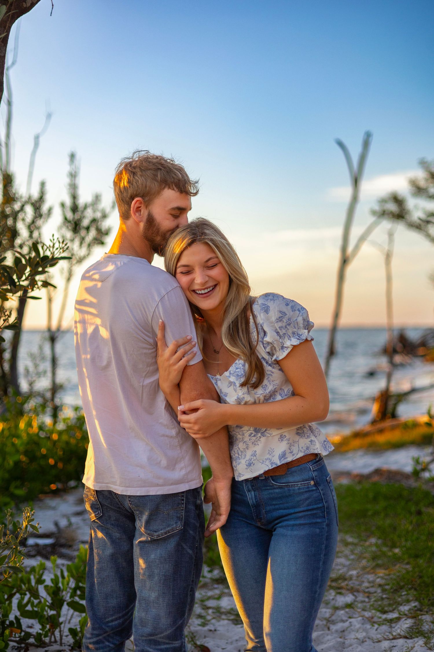 Couple laughing and embracing at sunset near a coastal beach with dead trees and golden hour light.