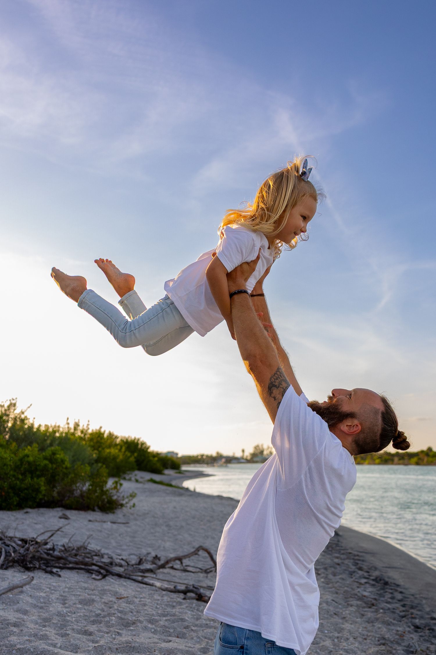 A father joyfully lifts his young blonde daughter high in the air on a sunny beach near calm water.