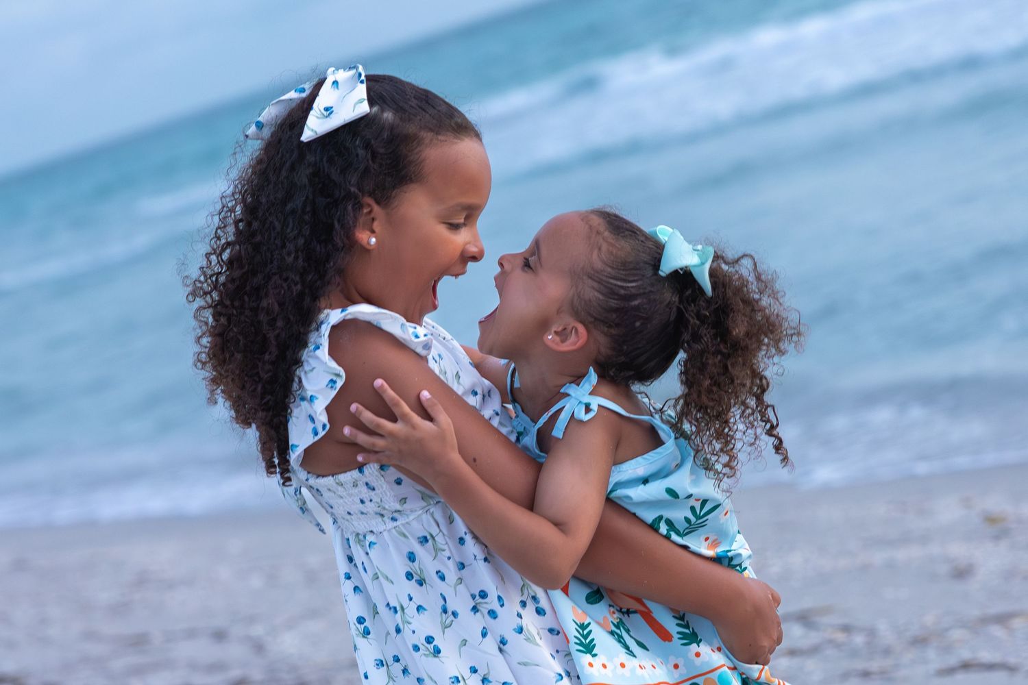 Two young girls in matching floral dresses embrace and smile at each other on a sandy beach.
